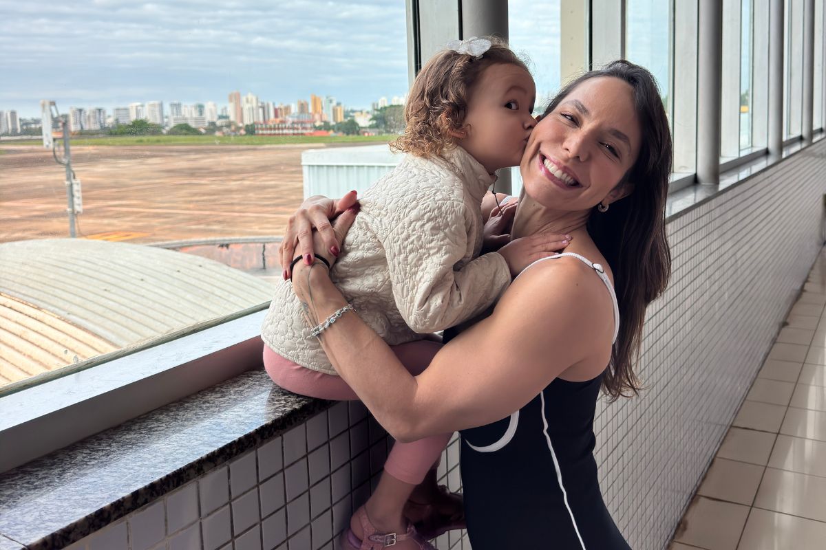 Mulher sorridente de cabelo castanho e blusa preta abraça uma menina de cabelo cacheado e casaco bege, que a beija na bochecha, em um aeroporto com vista para a cidade