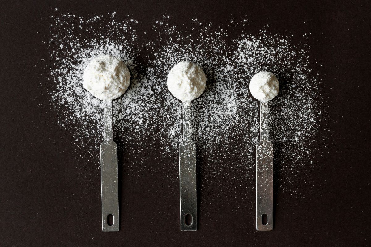 Três colheres de medida de metal, de tamanhos diferentes, alinhadas verticalmente em uma superfície escura. Cada colher contém uma porção de pó branco, que também está espalhado ao redor das colheres