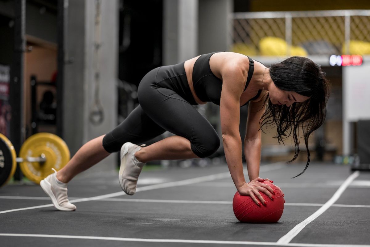 Mulher de cabelo escuro, vestindo top e calça legging pretos, tênis brancos, fazendo exercício de prancha com uma perna flexionada e as mãos apoiadas em uma bola vermelha no chão da academia