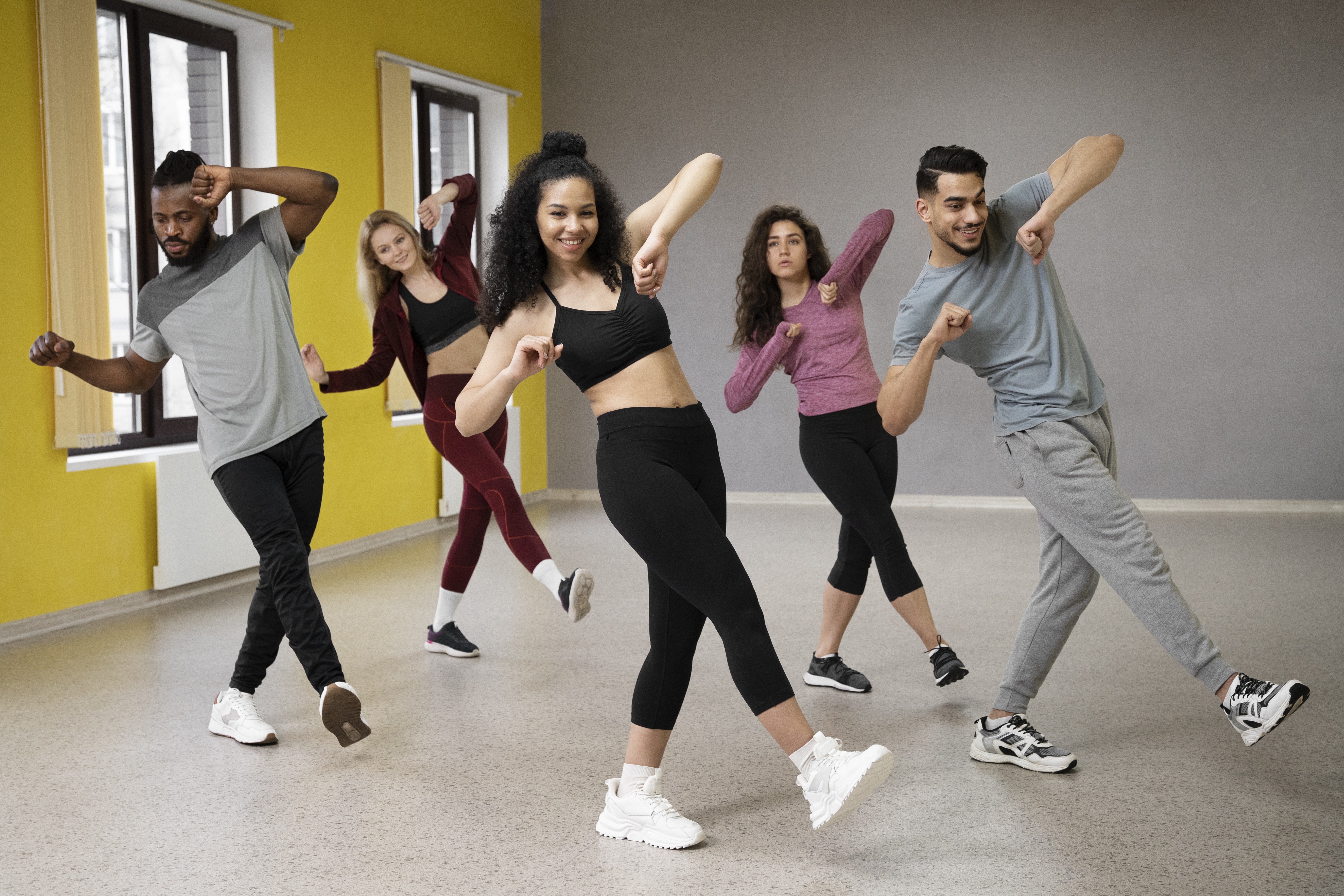 Cinco jovens adultos, três mulheres e dois homens, dançam em uma sala de aula de estúdio com paredes amarelas e cinzas. Todos sorriem e usam roupas esportivas, com braços e pernas flexionados em movimentos sincronizados