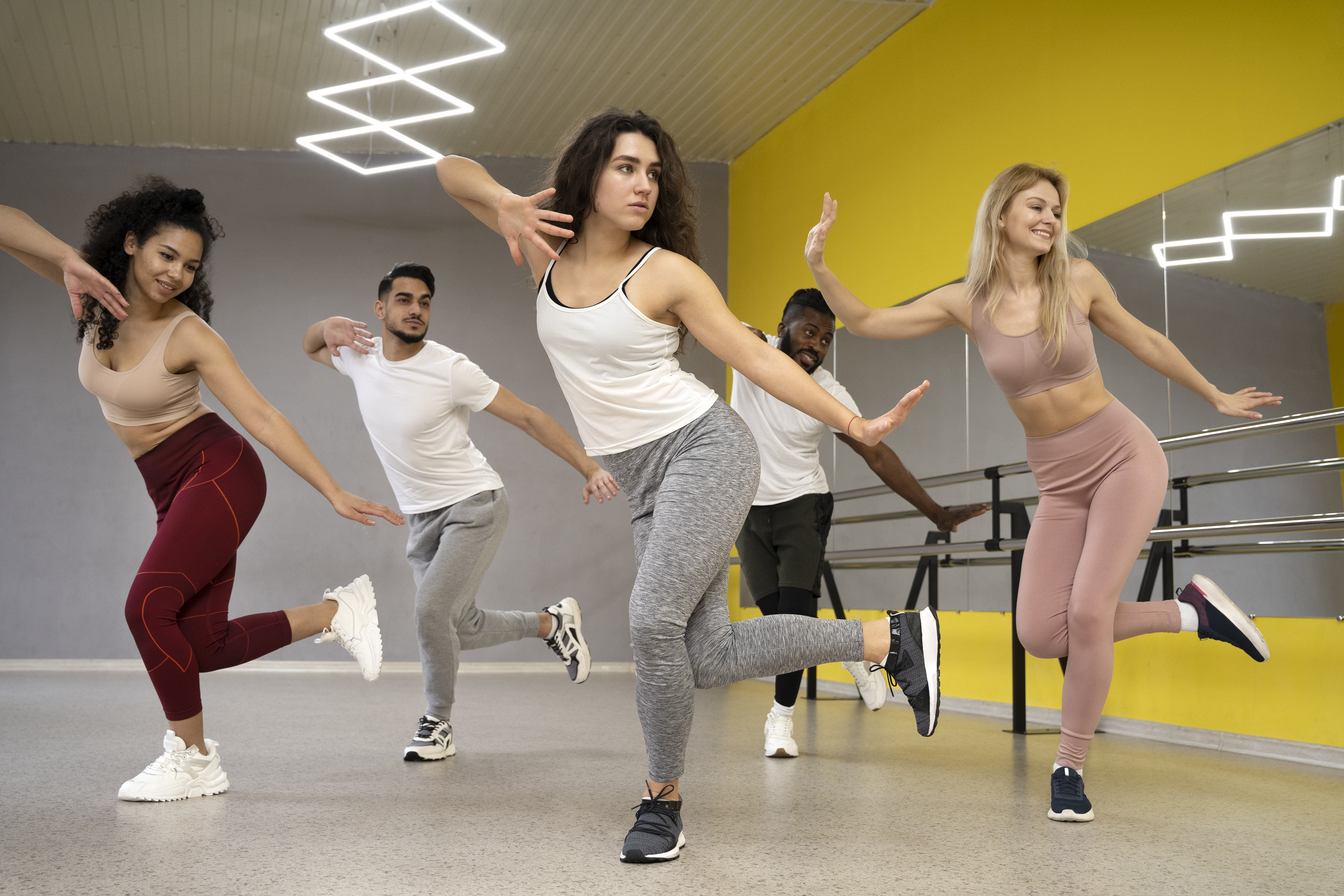 Cinco pessoas, tr&ecirc;s mulheres e dois homens, dan&ccedil;ando em uma sala de gin&aacute;stica com paredes cinzas e amarelas. Todos est&atilde;o com roupas esportivas e t&ecirc;nis, em uma pose de dan&ccedil;a com uma perna levantada e os bra&ccedil;os estendidos. A mulher do centro tem cabelos cacheados e veste regata branca e cal&ccedil;a cinza. A mulher da esquerda sorri, com top bege e cal&ccedil;a vinho. A mulher da direita sorri, com top e cal&ccedil;a rosa. Os homens, um com barba e outro negro, vestem camisetas brancas e cal&ccedil;as esportivas. H&aacute; um espelho na parede amarela e barras de bal&eacute; ao fundo