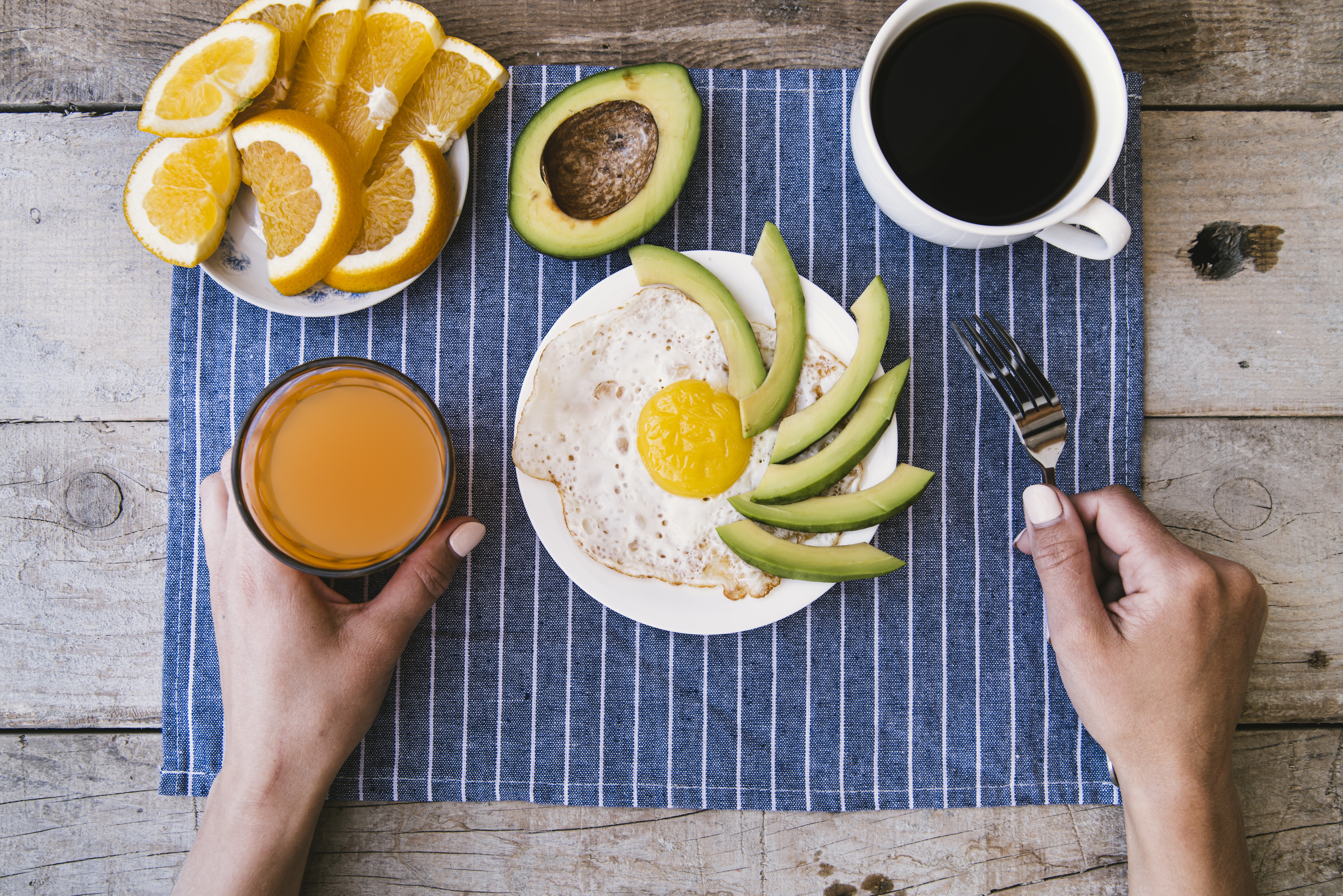 Duas mãos seguram um copo de suco de laranja e um garfo, sobre uma mesa de madeira rústica com um café da manhã saudável. Há um prato com ovo frito e fatias de abacate, um prato com fatias de laranja, um abacate cortado ao meio e uma xícara de café preto, tudo sobre um pano de prato azul listrado