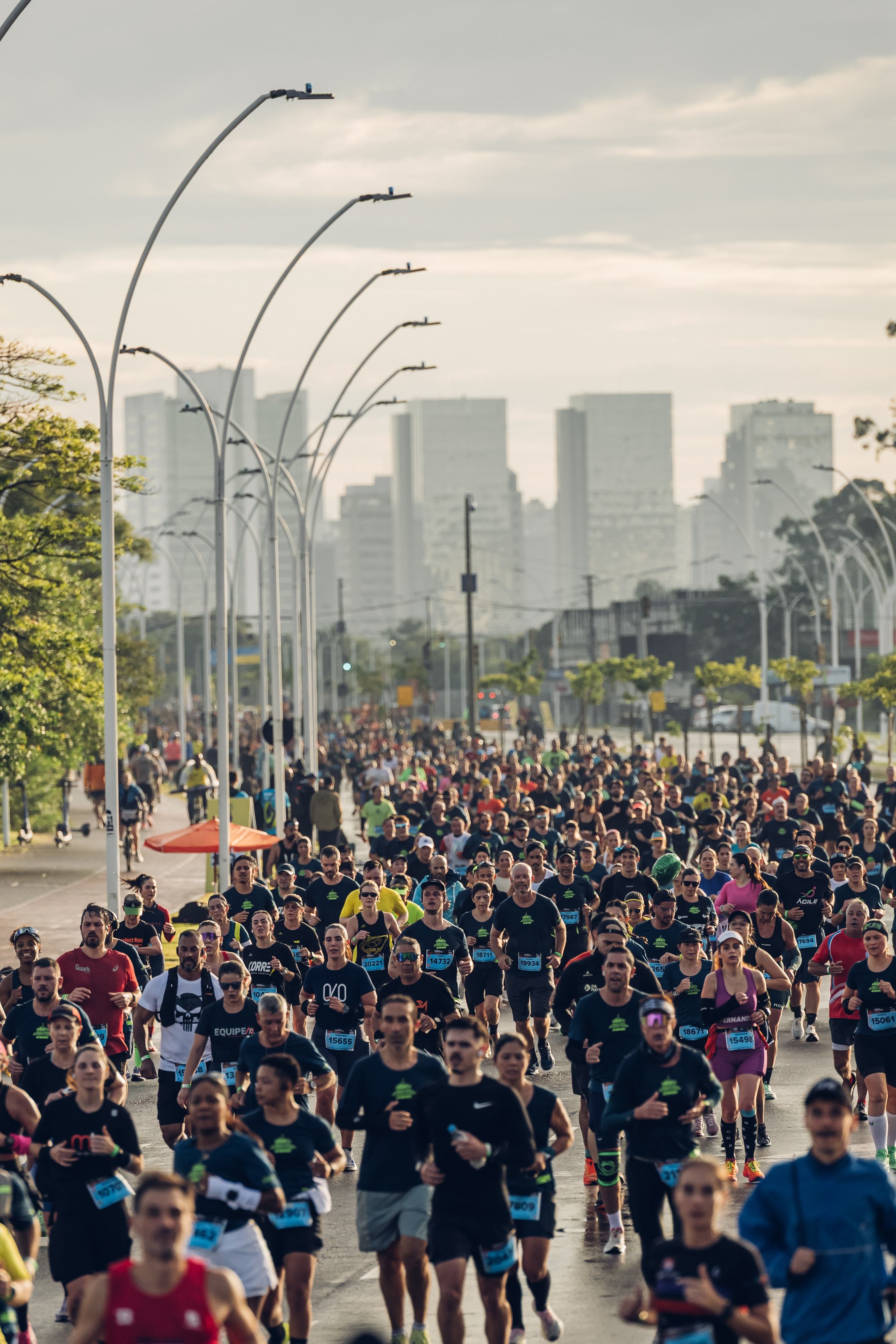 Multidão de corredores em uma maratona urbana, vestindo camisetas escuras e números de peito, em uma rua larga com postes de luz curvos e prédios altos ao fundo, sob céu nublado