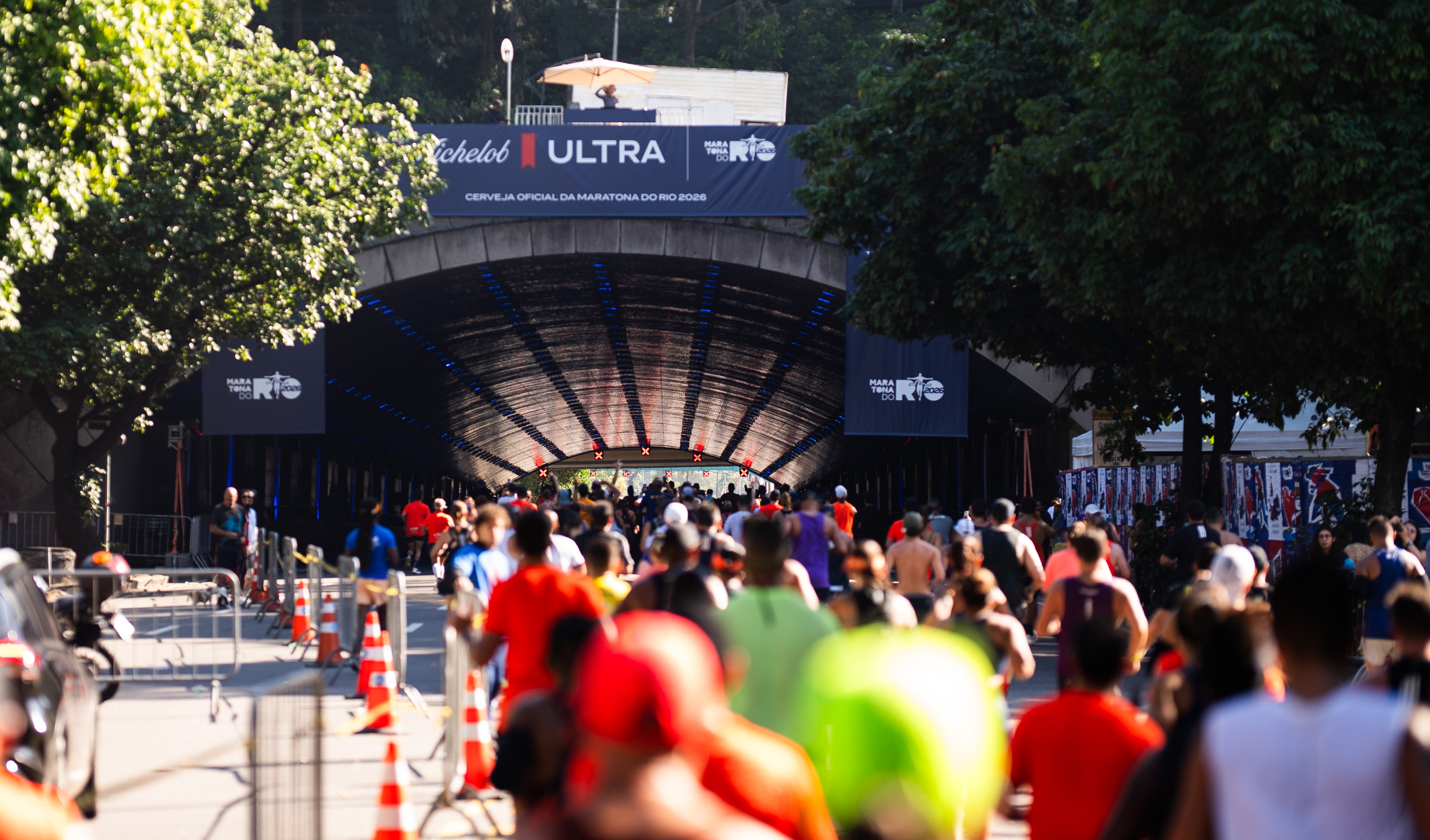 Corredores em uma maratona se aproximam de um túnel iluminado, com banners da Maratona do Rio 2026 e Michelob Ultra acima. A luz do sol incide sobre as árvores à esquerda, criando um contraste com a sombra do túnel. Cones de sinalização laranjas delimitam a pista.