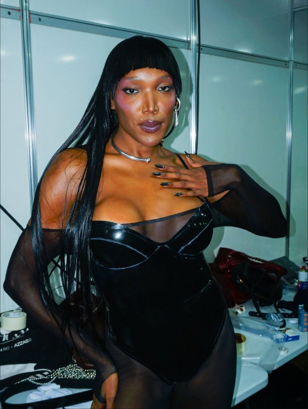 Pessoa de pele escura, cabelo liso preto com franja, usando body de látex preto e mangas transparentes, colar prateado e brincos longos. Ela tem maquiagem roxa nos olhos e lábios, unhas pretas e a mão esquerda no peito. O fundo é um camarim com uma mesa bagunçada