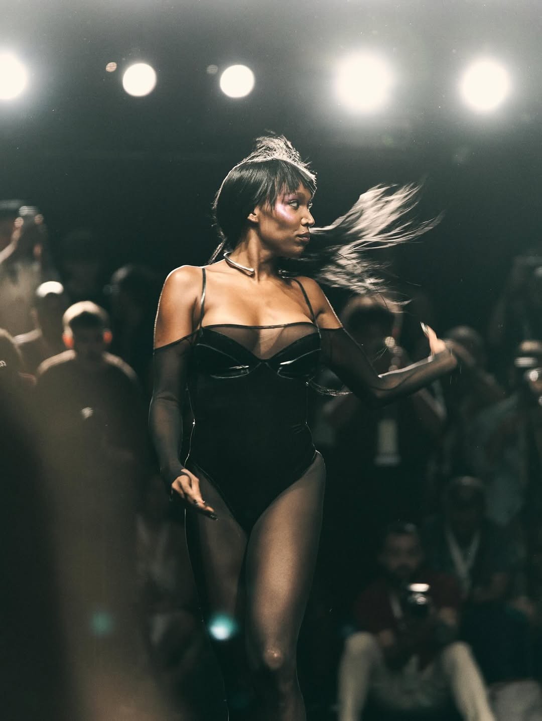 Modelo de pele escura com cabelo esvoaçante, usando body preto transparente e gargantilha prateada, desfilando em passarela escura com luzes fortes e público desfocado ao fundo