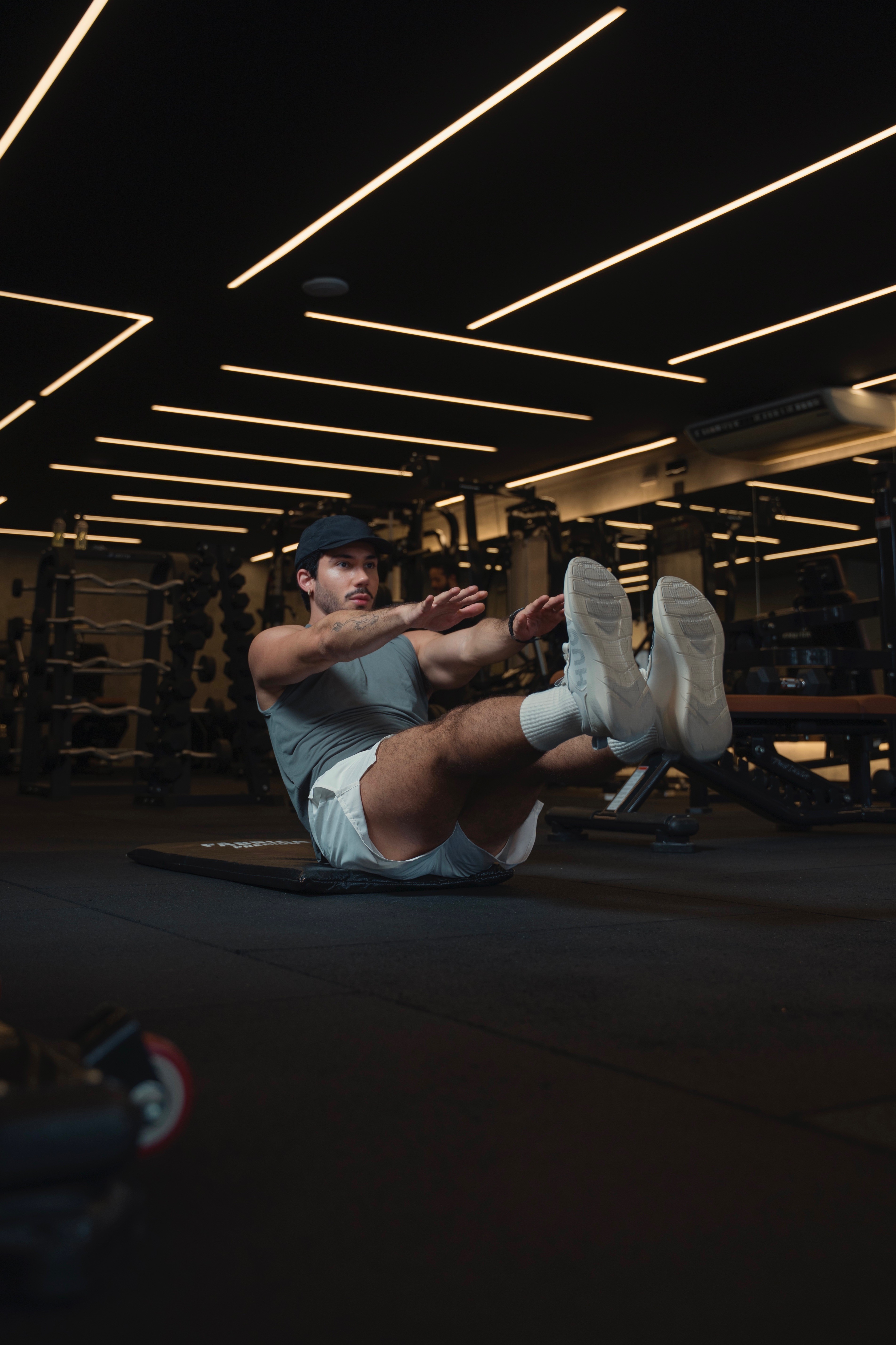 Homem jovem com bermuda e sapatos brancos, camiseta cinza e usando boné em uma academia, fazendo abdominal com os braços esticados.