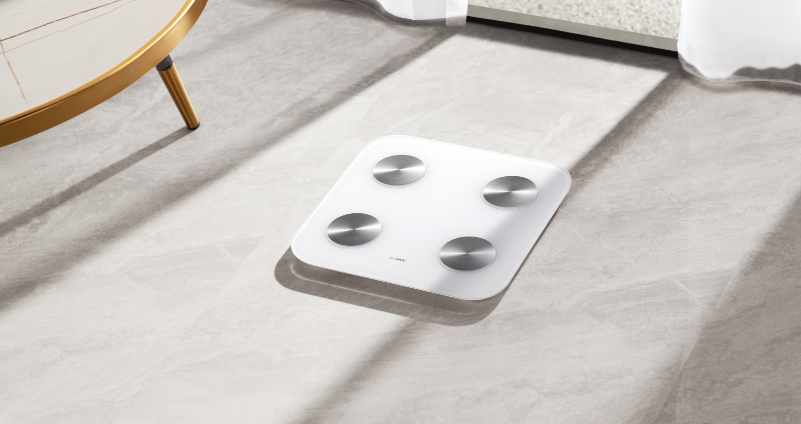 Balança inteligente Huawei branca com quatro sensores metálicos redondos, sobre piso de mármore claro, próxima a uma mesa lateral dourada e uma janela com cortina branca