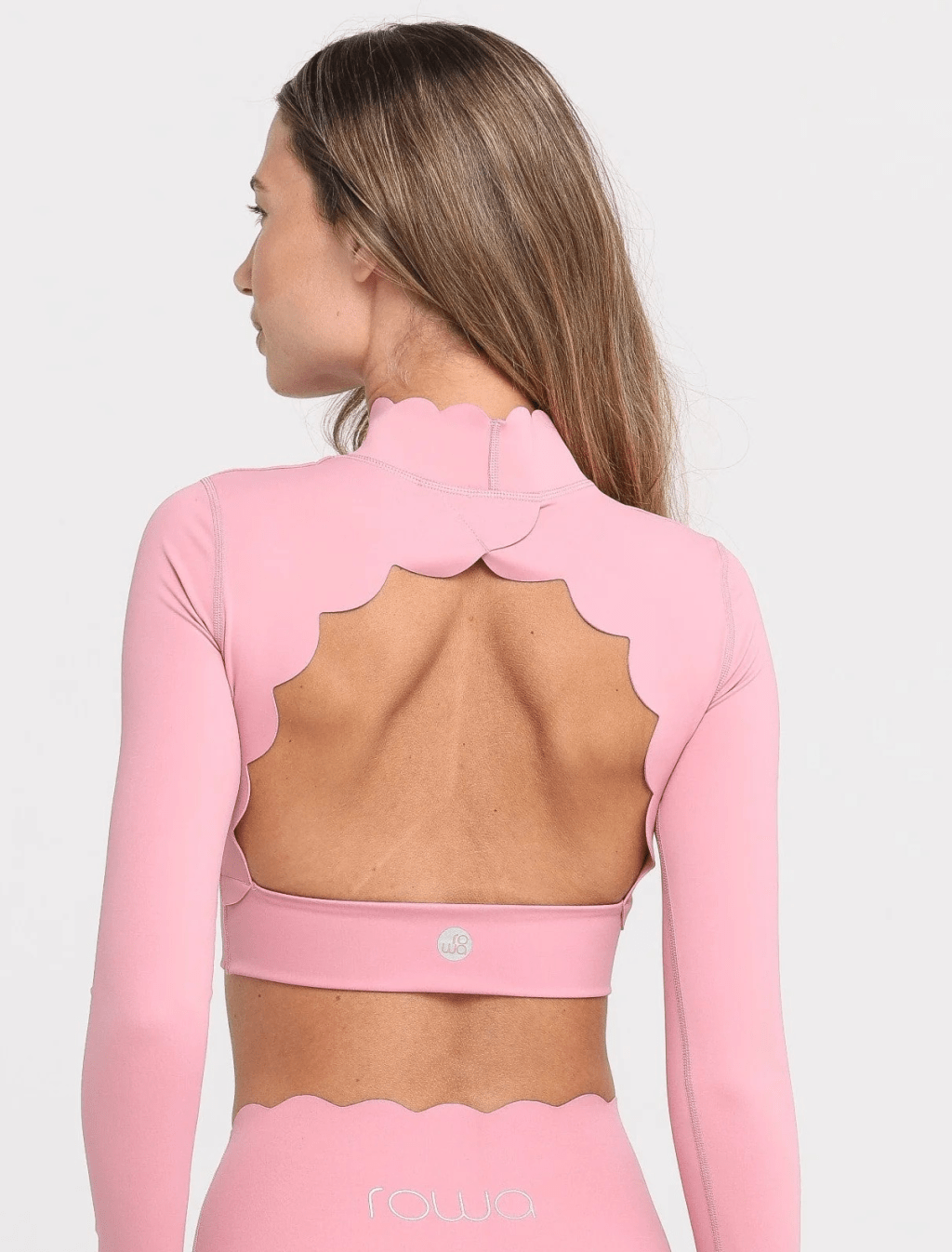 Mulher de costas, com cabelos castanhos longos, vestindo um conjunto esportivo rosa claro. A blusa de manga comprida tem gola alta e recortes ondulados nas costas, expondo a parte superior das costas. A calça tem cintura alta e acabamento ondulado na borda superior. O logo rowa aparece na calça e um símbolo circular na blusa