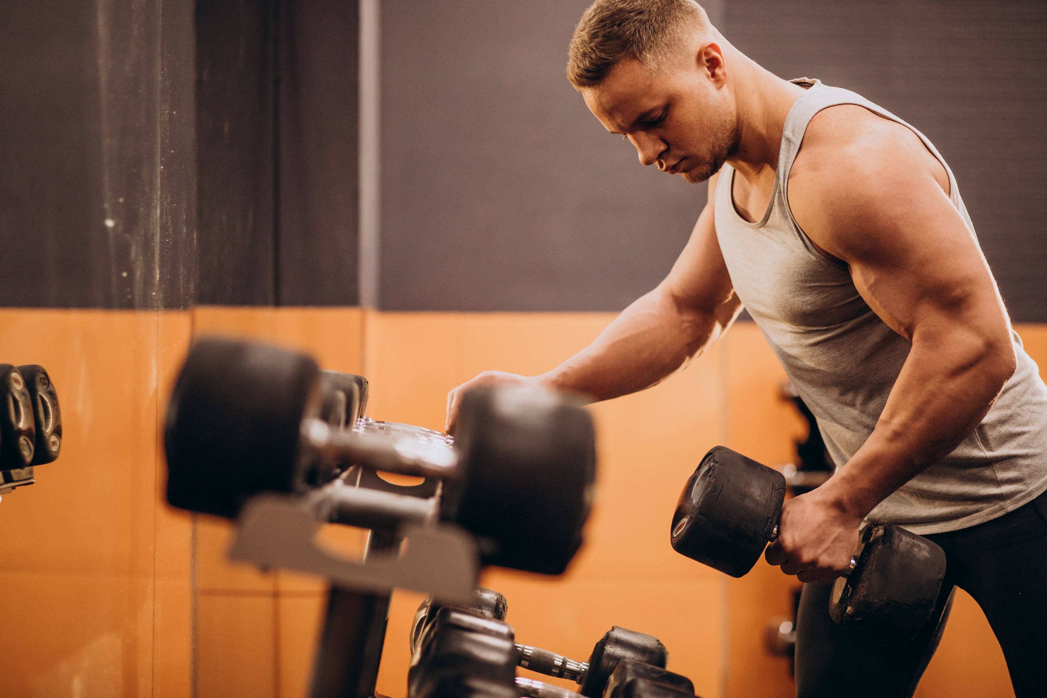 Homem branco musculoso, de perfil, com regata cinza e calça preta, pegando um halter preto com a mão direita em uma academia com parede laranja e preta