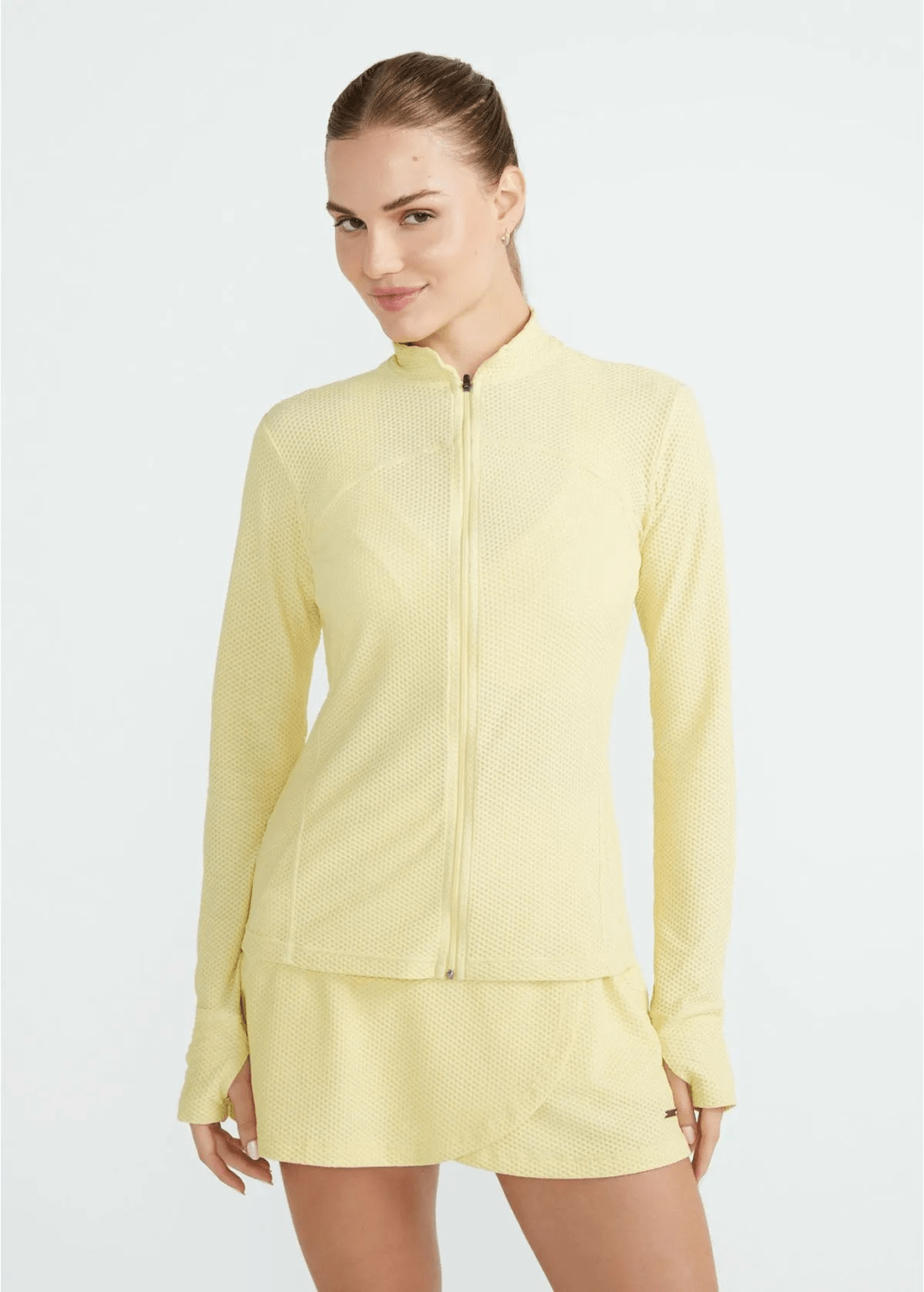 Mulher jovem, de pele clara e cabelo castanho preso, sorri levemente enquanto veste um conjunto esportivo amarelo-claro texturizado, composto por jaqueta de zíper e saia-short, em fundo branco