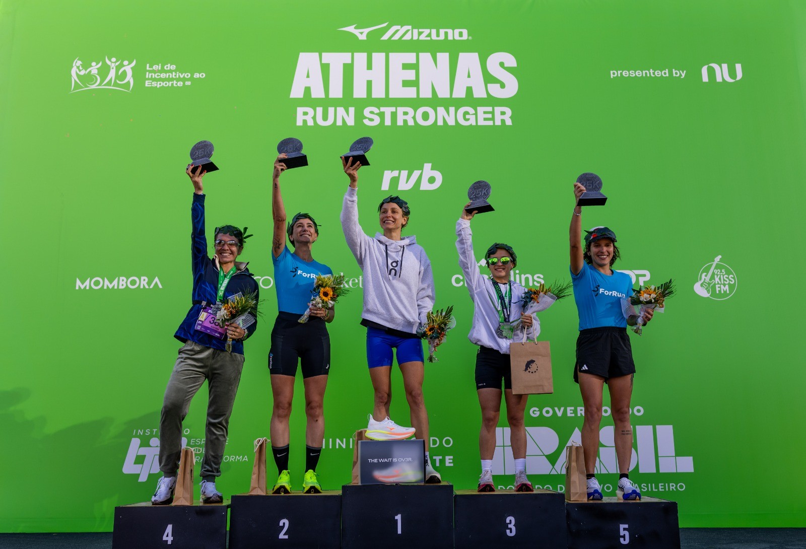 Cinco mulheres atletas sorridentes no pódio de uma corrida, levantando troféus e segurando buquês de girassóis, em frente a um painel verde com logos de patrocinadores