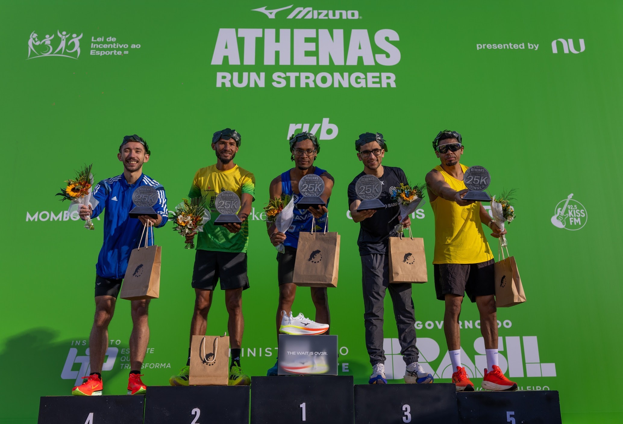 Cinco homens sorridentes, usando coroas de louros, em um pódio de corrida. O primeiro lugar exibe um tênis branco, os outros seguram troféus, buquês e sacolas de papel. O fundo verde tem o logo ATHENAS RUN STRONGER e patrocinadores