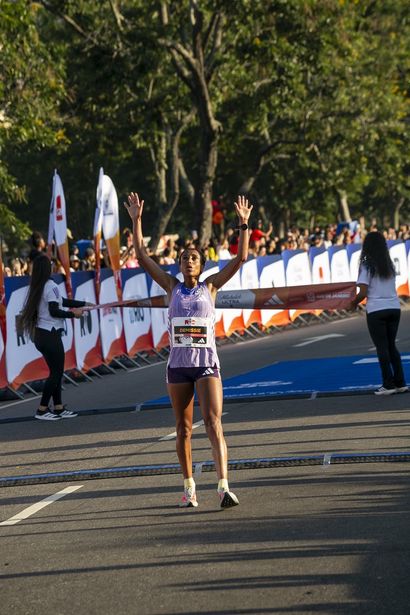 Corredora de pele escura, com camiseta e shorts roxos, braços erguidos em celebração ao cruzar a linha de chegada de uma maratona. Ao fundo, público e árvores verdes sob luz solar