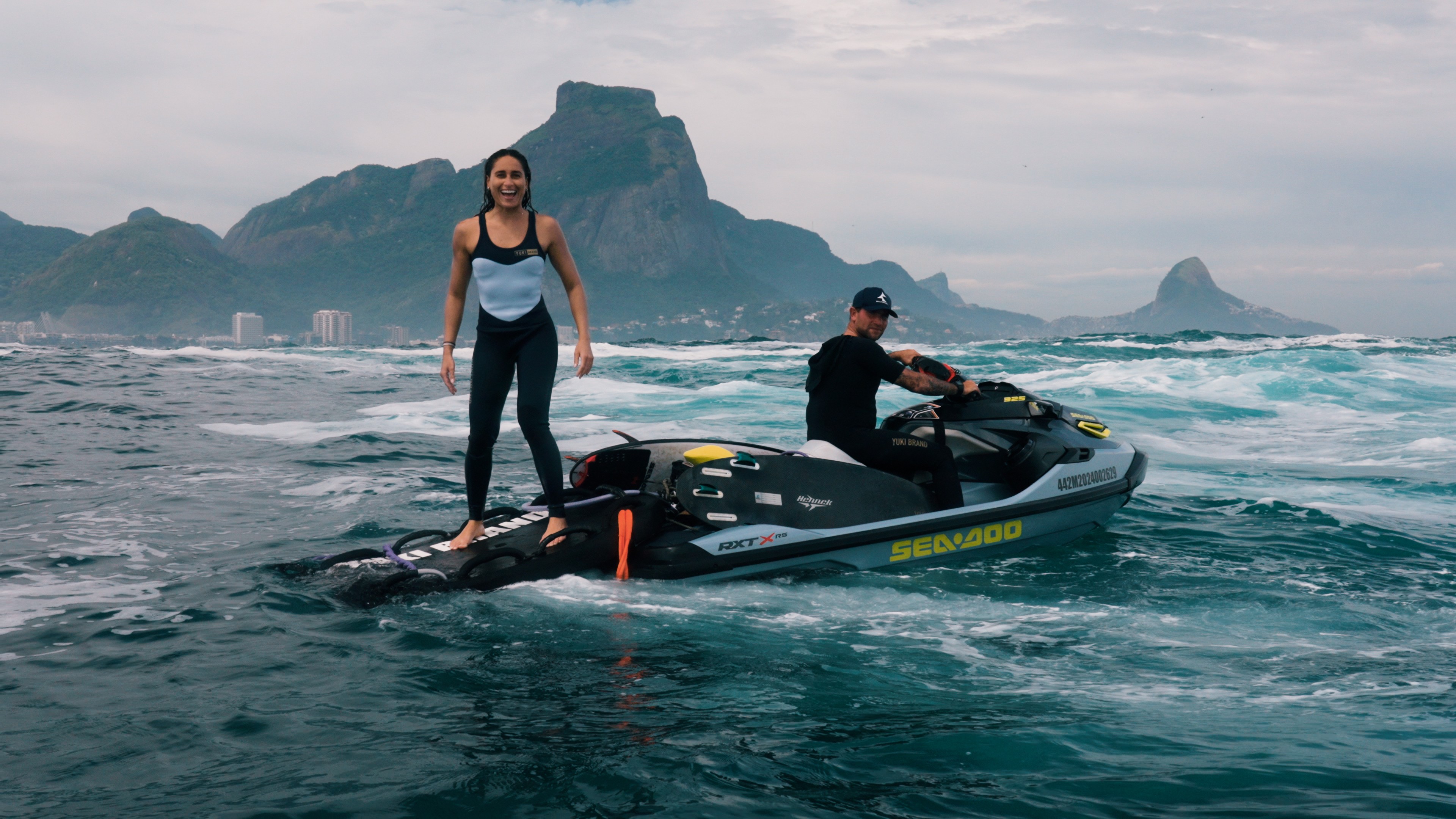 Mulher sorridente em roupa de neoprene preta e azul claro, de pé sobre uma prancha de surf rebocada por um jet ski cinza pilotado por um homem de boné e roupa preta, em mar agitado com montanhas e prédios ao fundo