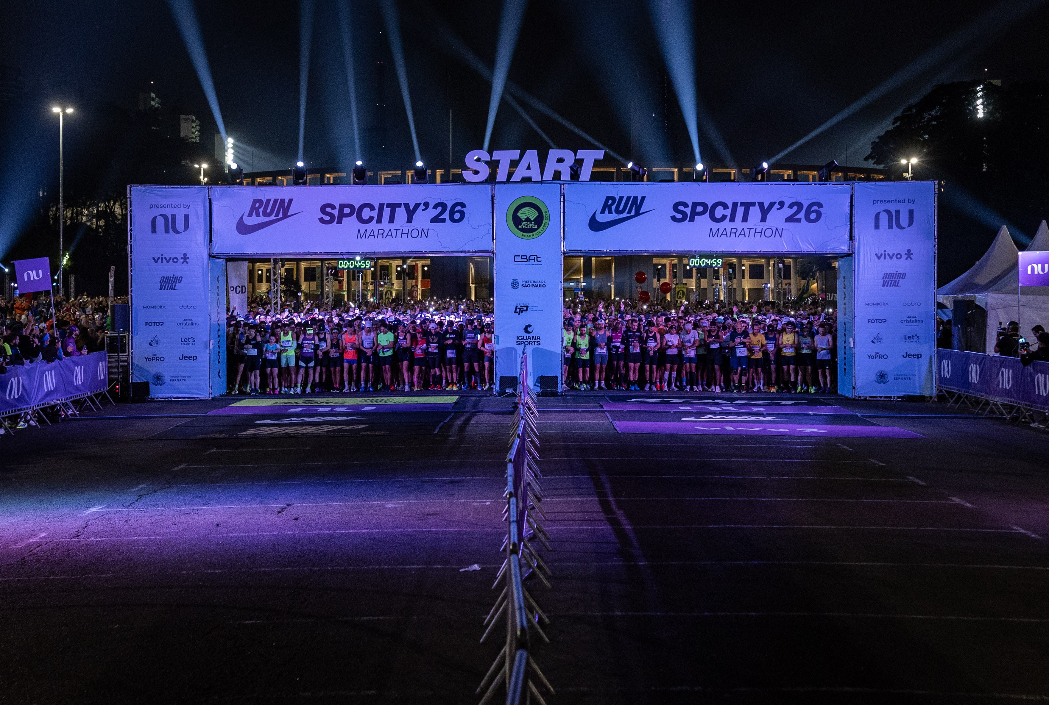 Corredores aguardam o início da SP City Marathon 26 sob um portal START iluminado por holofotes, com banners de patrocinadores e um cronômetro digital exibindo 00:04:59. A multidão, vestindo roupas esportivas, está alinhada na rua escura, que reflete a iluminação roxa do evento.