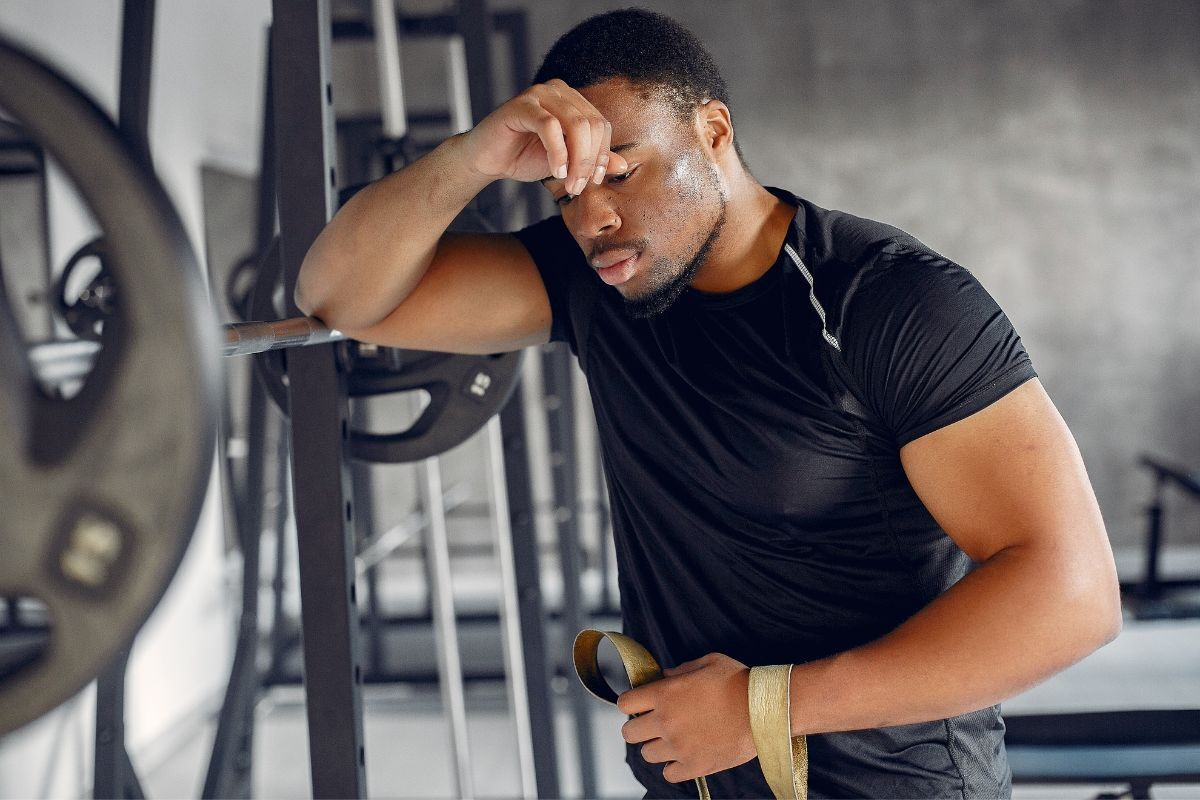 Homem negro, com barba rala e camiseta preta, suado, encosta a cabeça na mão e o cotovelo em uma barra de peso na academia, segurando uma faixa de levantamento de peso amarela