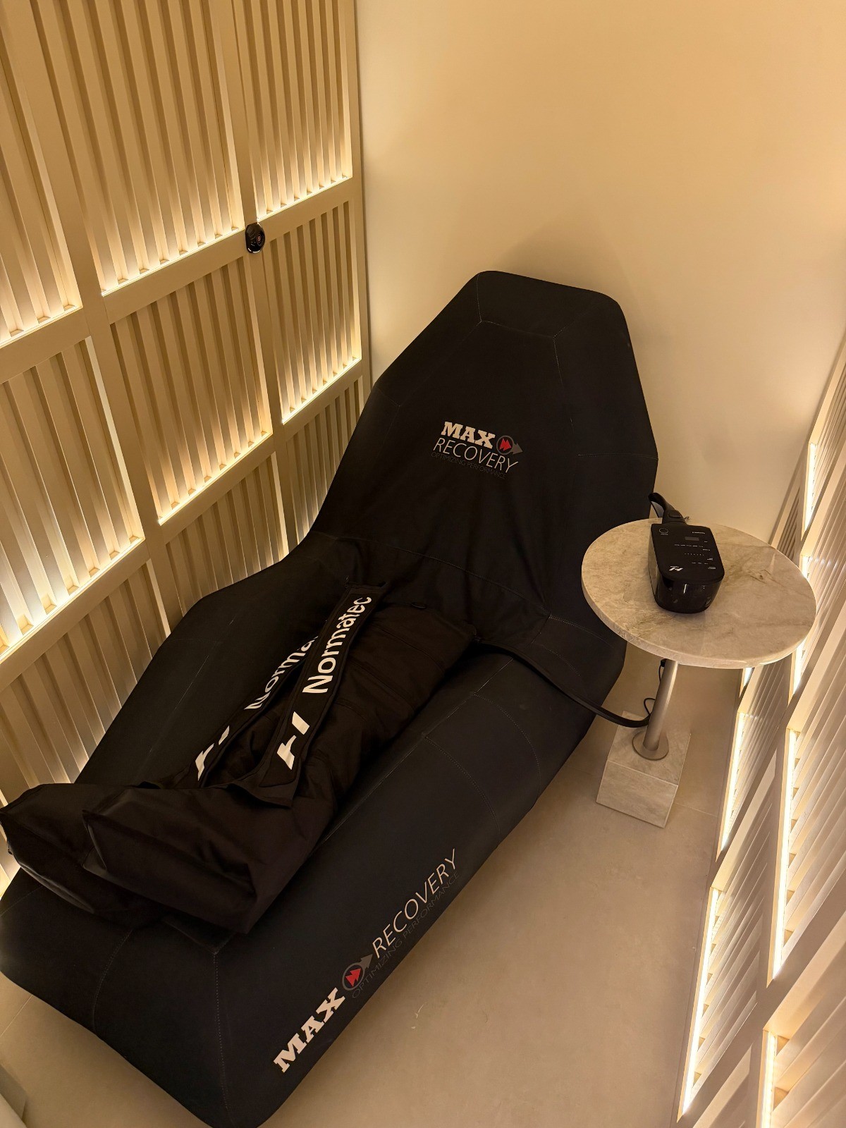Poltrona reclinável preta da marca Max Recovery com botas de compressão Normatec, ao lado de uma mesa de cabeceira redonda de mármore com um aparelho eletrônico preto, em um ambiente com paredes de ripas de madeira iluminadas por luzes indiretas