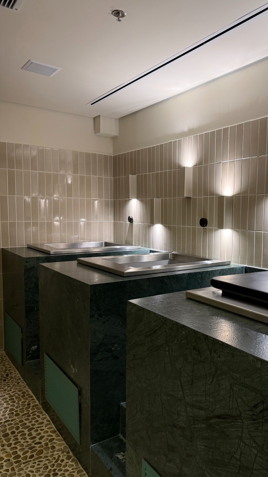 Ambiente interno com três bancadas de mármore verde escuro e cubas de inox. A parede é revestida com azulejos bege claros e há luminárias de parede que emitem luz para cima e para baixo. O chão é de pedras claras e o teto tem iluminação linear e um sprinkler.