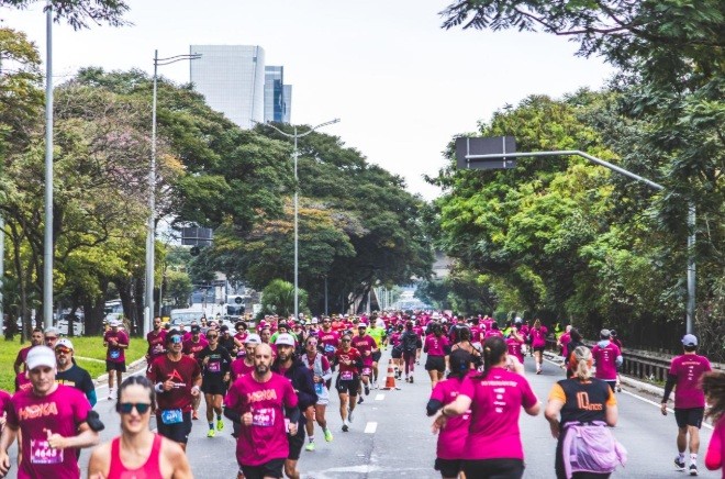 Multidão de corredores, a maioria vestindo camisetas magenta, participando de uma corrida de rua em uma avenida arborizada com prédios ao fundo