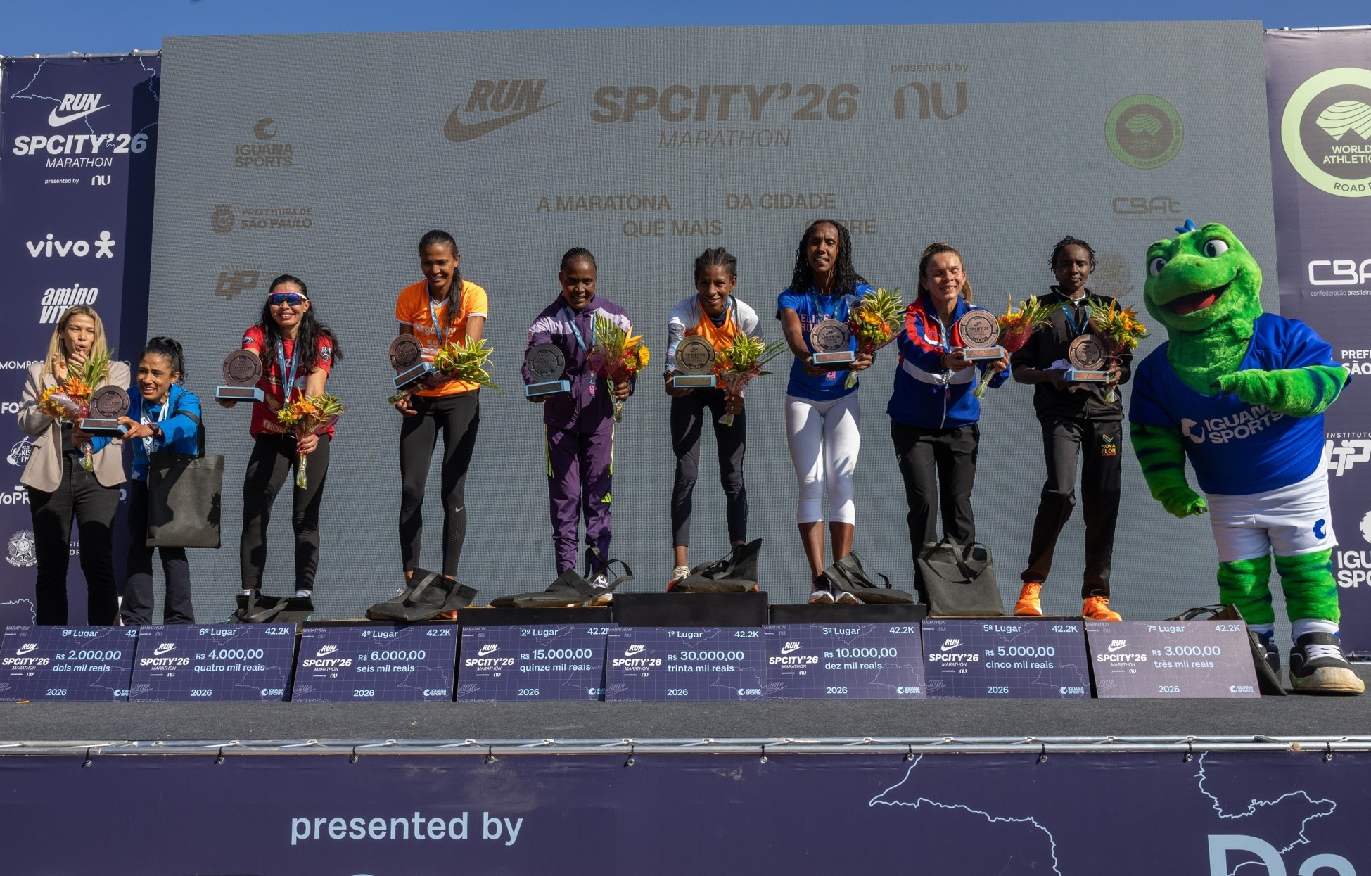 Oito mulheres e um mascote verde em um pódio de premiação de maratona. As mulheres seguram troféus e buquês, vestindo roupas esportivas coloridas. O mascote, um lagarto verde com camiseta azul Iguana Sports, está à direita. No fundo, um telão exibe logotipos de patrocinadores e informações da SPCITY26 Marathon. Abaixo do pódio, placas indicam os lugares, valores de premiação e distância de 42.2K