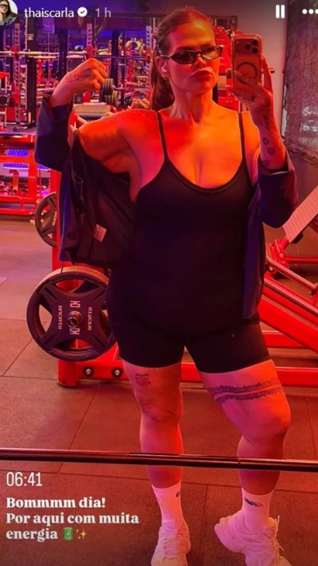 Thais Carla posa em selfie de corpo inteiro na academia, vestindo top e shorts pretos, jaqueta azul escura e óculos de sol. Ela faz bico e flexiona o braço direito, exibindo tatuagens nos braços e pernas. O ambiente tem iluminação avermelhada e equipamentos de musculação vermelhos ao fundo. No canto inferior esquerdo, texto branco diz 06:41 Bommmm dia! Por aqui com muita energia.