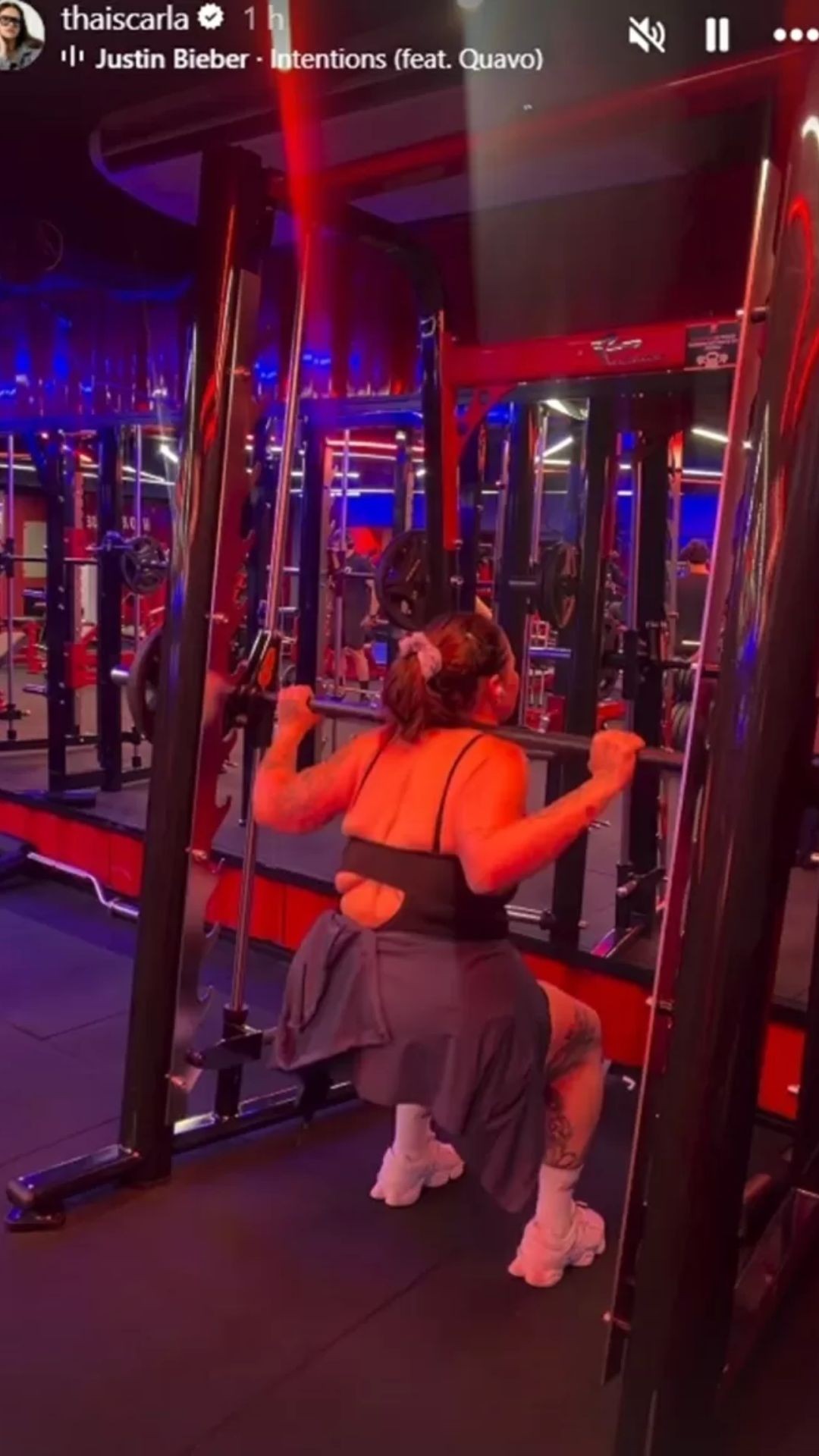 Mulher de costas, com top preto e calça cinza, agacha segurando uma barra de peso em uma máquina de academia com iluminação vermelha e azul