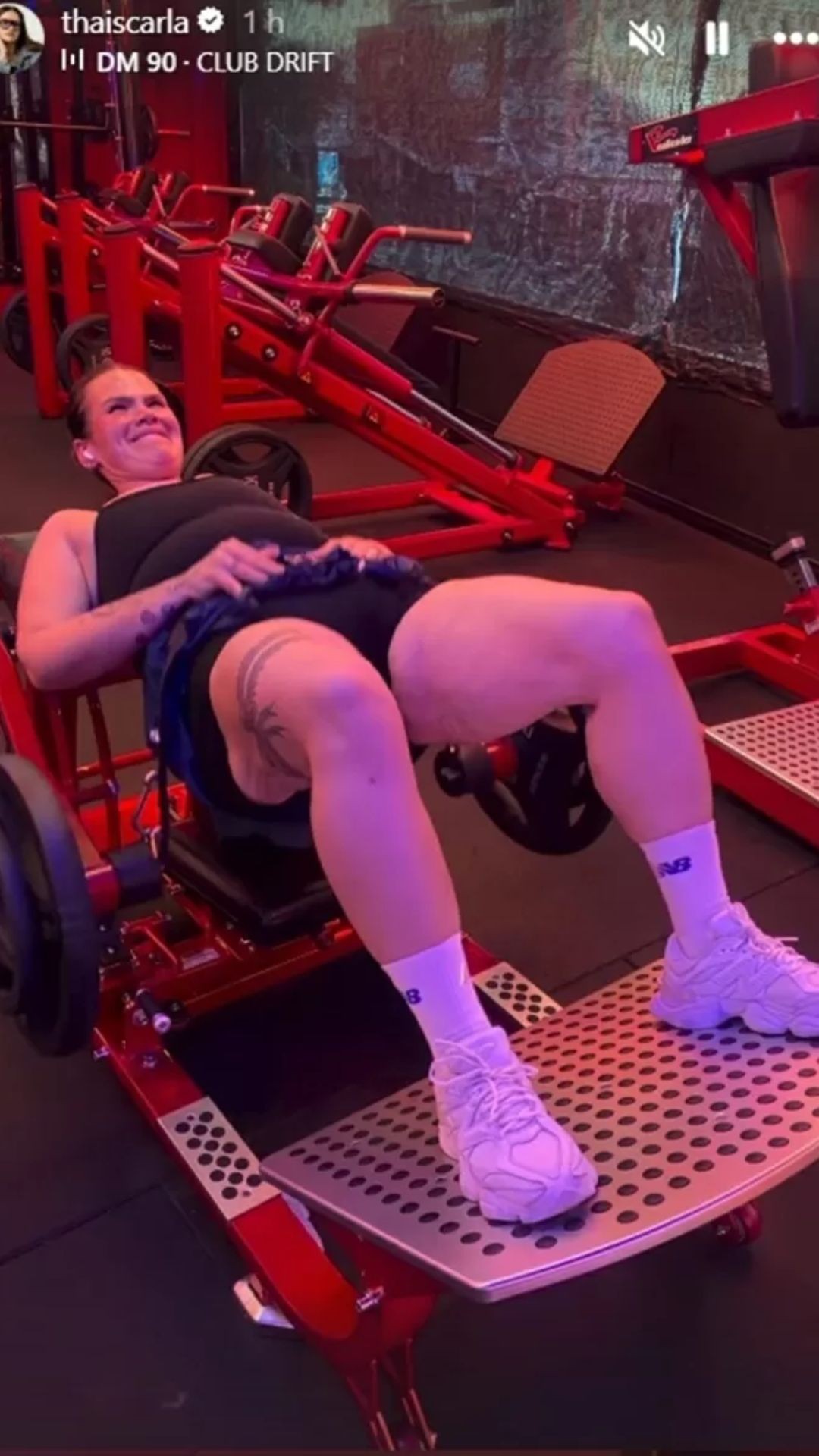 Mulher deitada em aparelho de academia vermelho, fazendo exercício para as pernas, com expressão de esforço e sorriso. Veste top preto, shorts escuro e tênis brancos, exibindo tatuagens na coxa.
