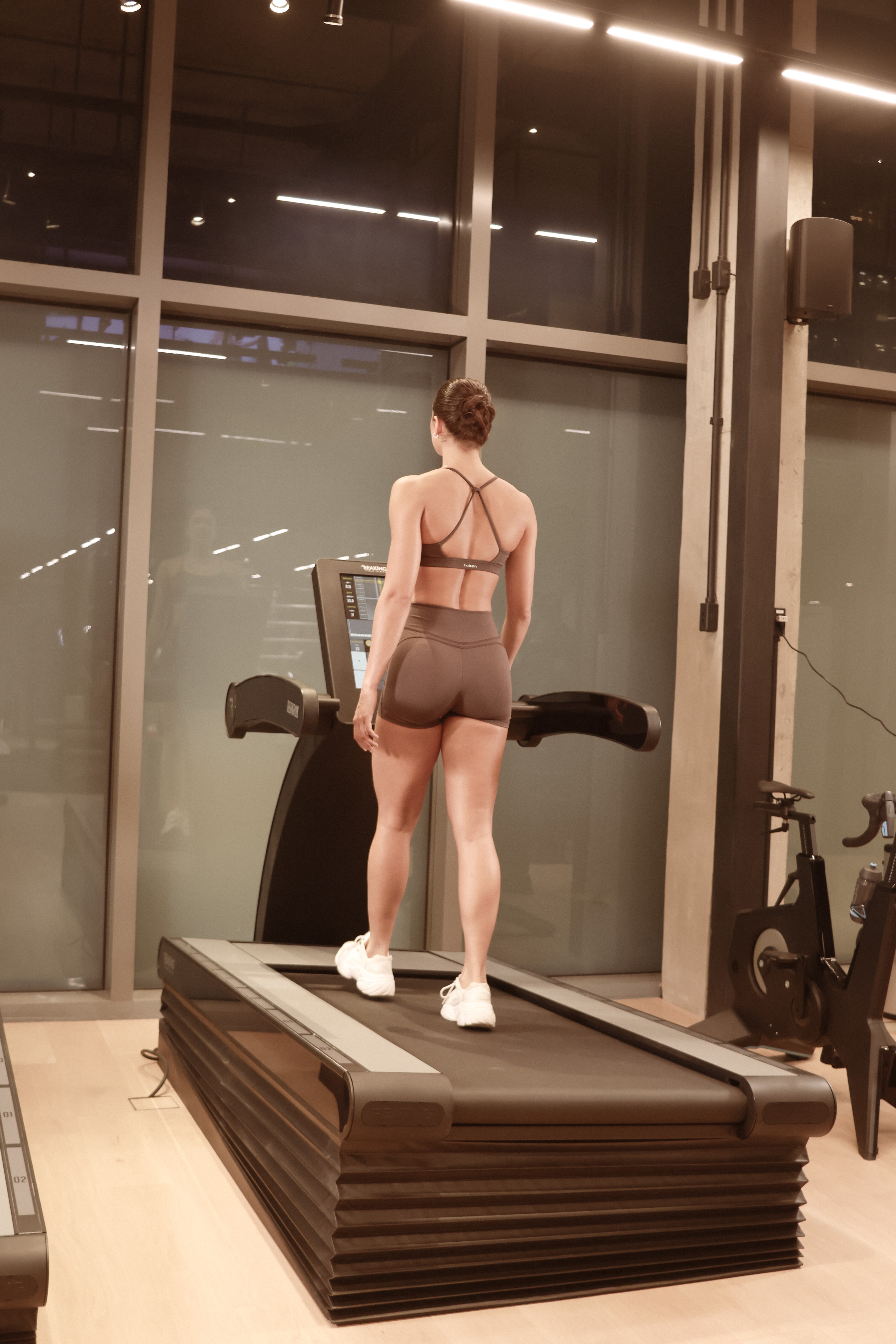 Mulher de costas, com coque e roupas esportivas marrons, caminha em esteira moderna dentro de uma academia com janelas grandes e luzes brancas no teto