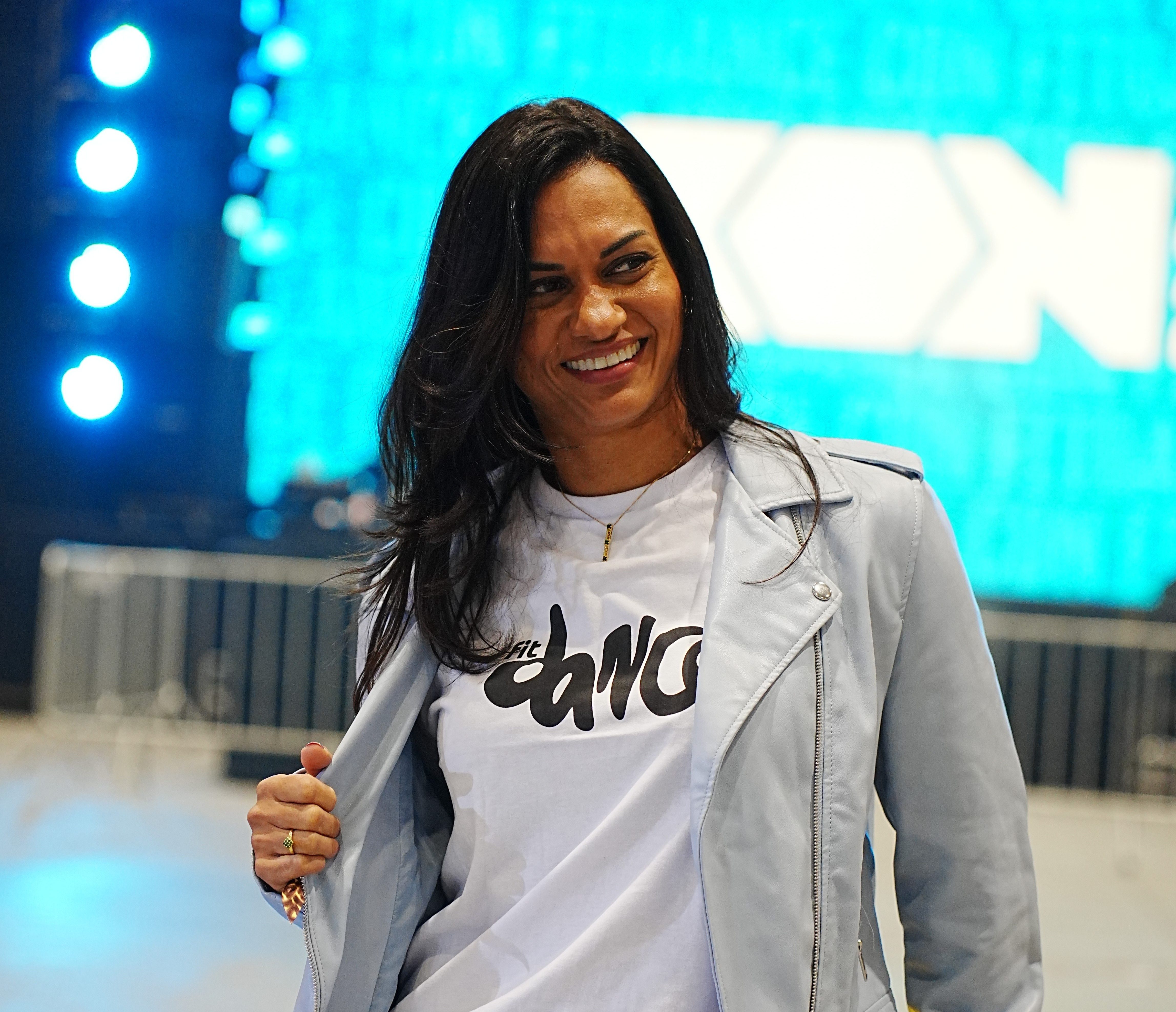 Mulher de pele morena e cabelos escuros, sorrindo, vestindo jaqueta azul-clara, camiseta branca com estampa preta e cal&ccedil;a jeans, sentada em superf&iacute;cie amarela, com fundo azul e luzes