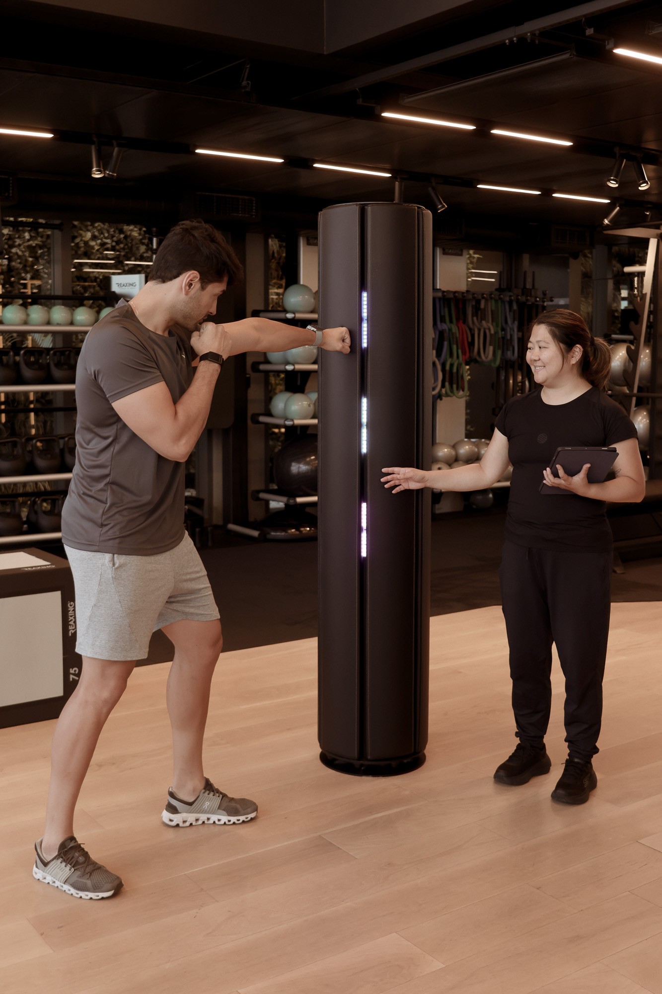 Homem socando um aparelho de treino vertical com luzes, enquanto uma mulher sorridente, segurando um tablet, observa e aponta para o aparelho em uma academia moderna