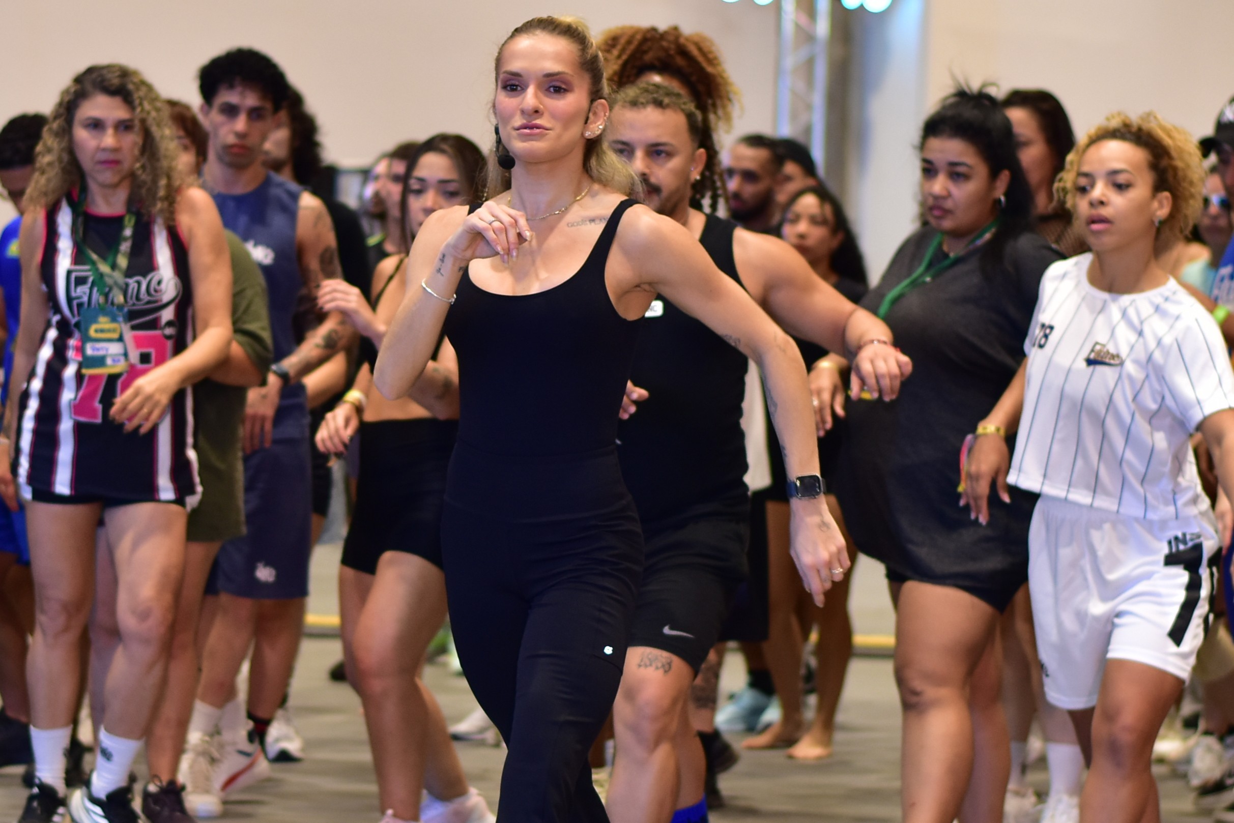 Grupo de pessoas em um evento, com uma mulher loira de top preto em primeiro plano, bra&ccedil;os flexionados e olhar concentrado. Ao fundo, outras pessoas de diversas etnias e idades, algumas usando roupas esportivas, participam da mesma atividade, possivelmente uma aula de dan&ccedil;a ou exerc&iacute;cio f&iacute;sico. O ambiente &eacute; amplo e iluminado, com foco na intera&ccedil;&atilde;o e movimento dos participantes