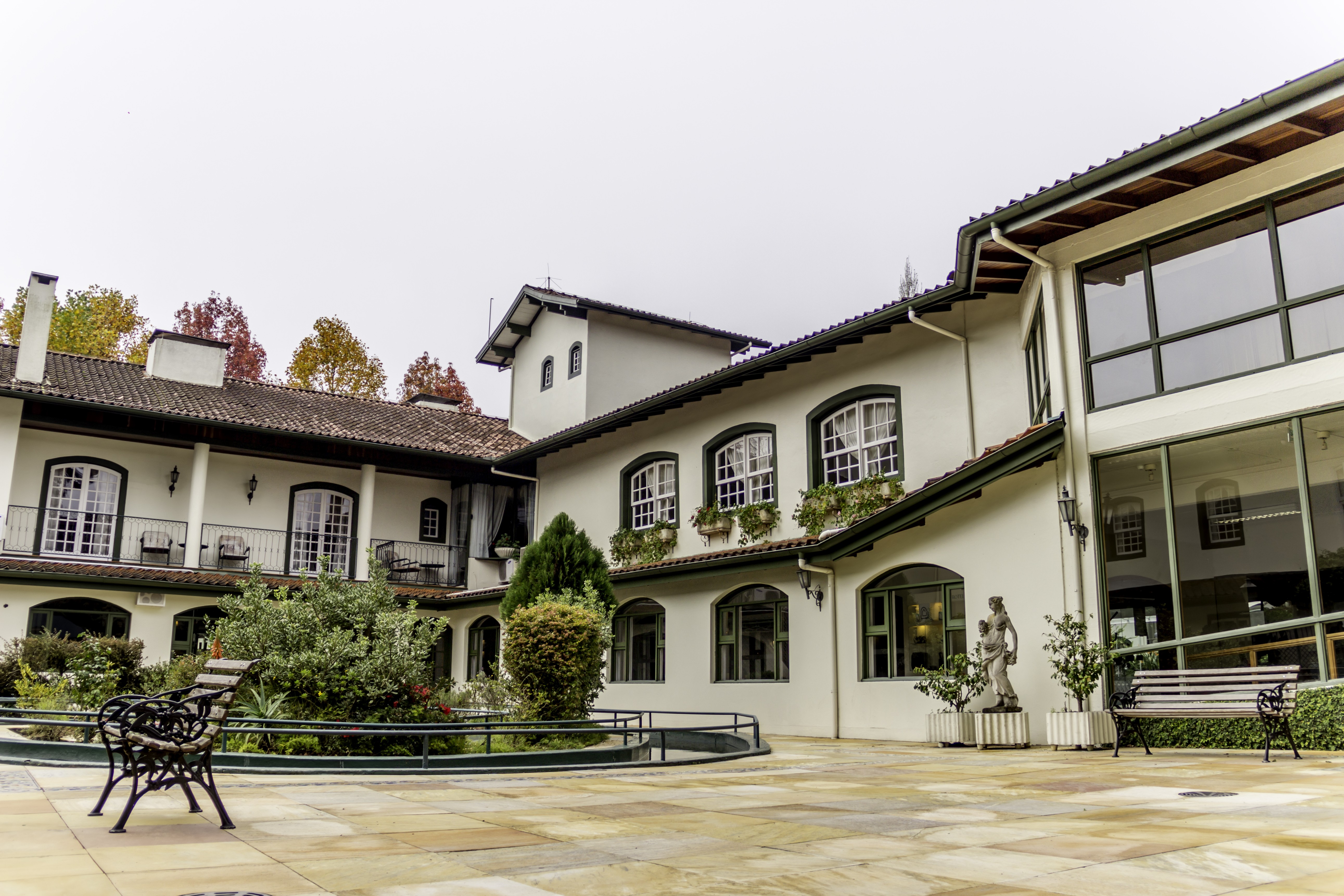 P&aacute;tio externo de um edif&iacute;cio branco com telhado de telhas marrons, janelas em arco e retangulares, e varandas com grades pretas. H&aacute; bancos de ferro e madeira, arbustos e &aacute;rvores com folhagem colorida de outono ao fundo, sob um c&eacute;u nublado