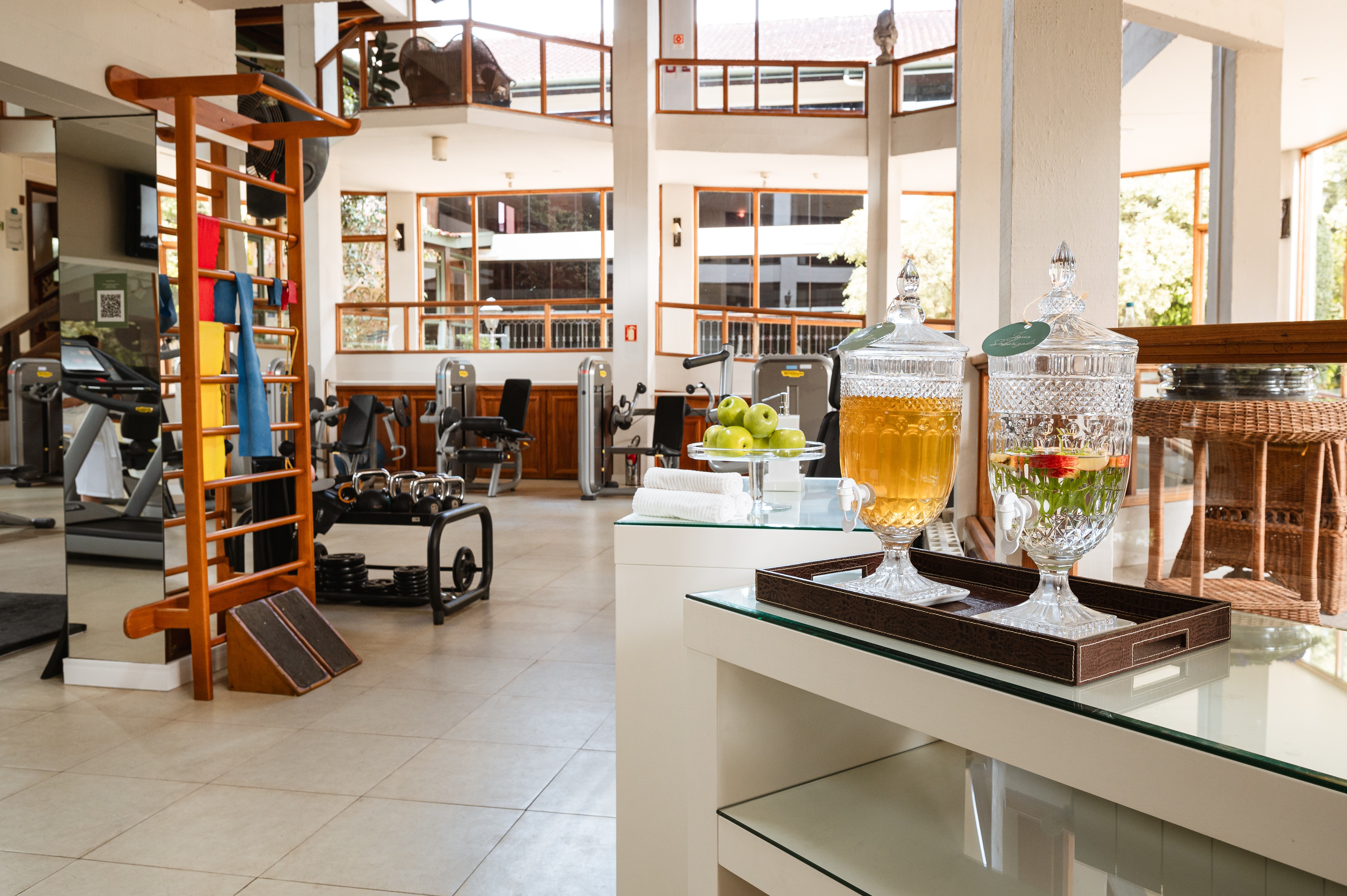 Academia espa&ccedil;osa e iluminada com equipamentos de muscula&ccedil;&atilde;o e esteiras. Em primeiro plano, dois dispensadores de bebidas de vidro, um com ch&aacute; gelado e outro com &aacute;gua saborizada com frutas, sobre uma bandeja escura. Ma&ccedil;&atilde;s verdes e toalhas brancas dobradas est&atilde;o em uma mesa branca ao lado
