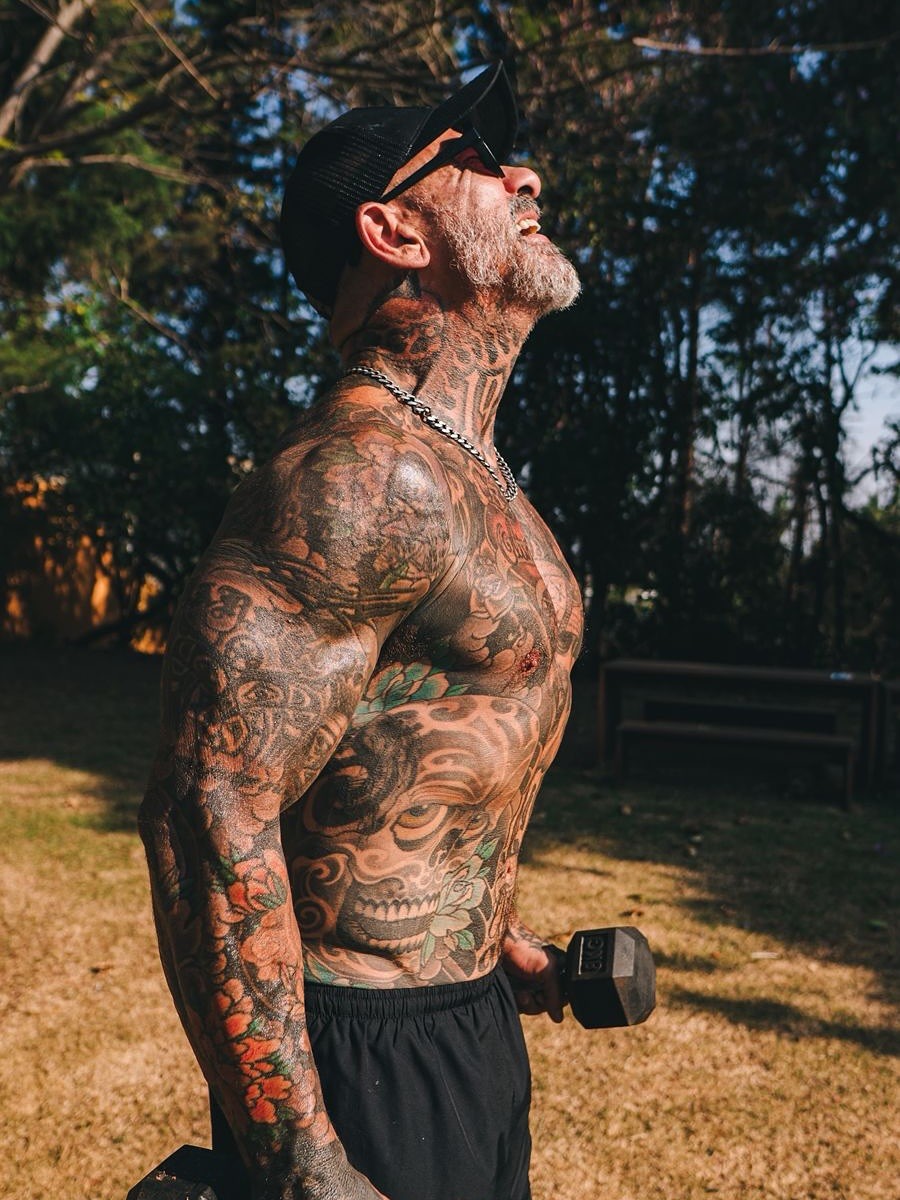 Homem musculoso e tatuado, com barba grisalha, boné preto e óculos escuros, levanta o rosto para o céu enquanto segura um halter em cada mão, exercitando-se ao ar livre em um dia ensolarado