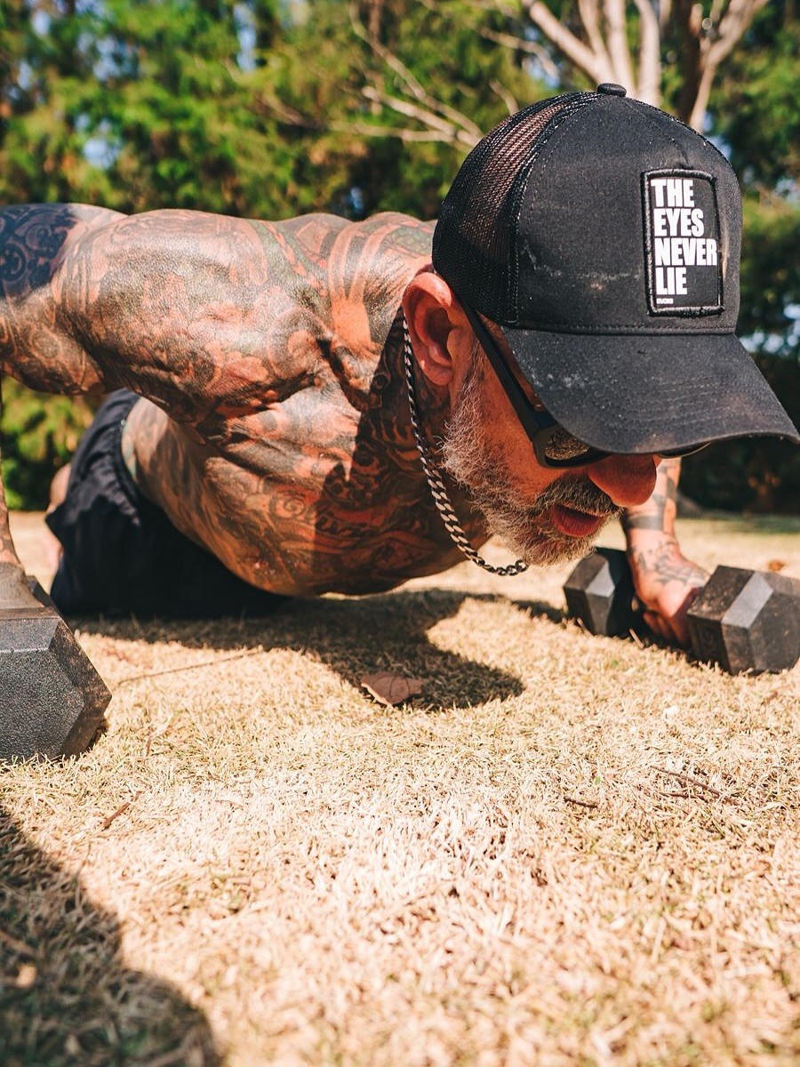 Homem musculoso e tatuado, de bon&eacute; preto e &oacute;culos escuros, fazendo flex&atilde;o com halteres na grama seca, sob luz solar intensa