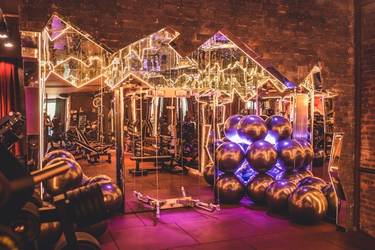 Academia moderna com paredes de tijolos e espelhos angulares iluminados por luzes neon amarelas. O ch&atilde;o &eacute; roxo e h&aacute; equipamentos de muscula&ccedil;&atilde;o, halteres e pilhas de bolas de gin&aacute;stica escuras com reflexos roxos