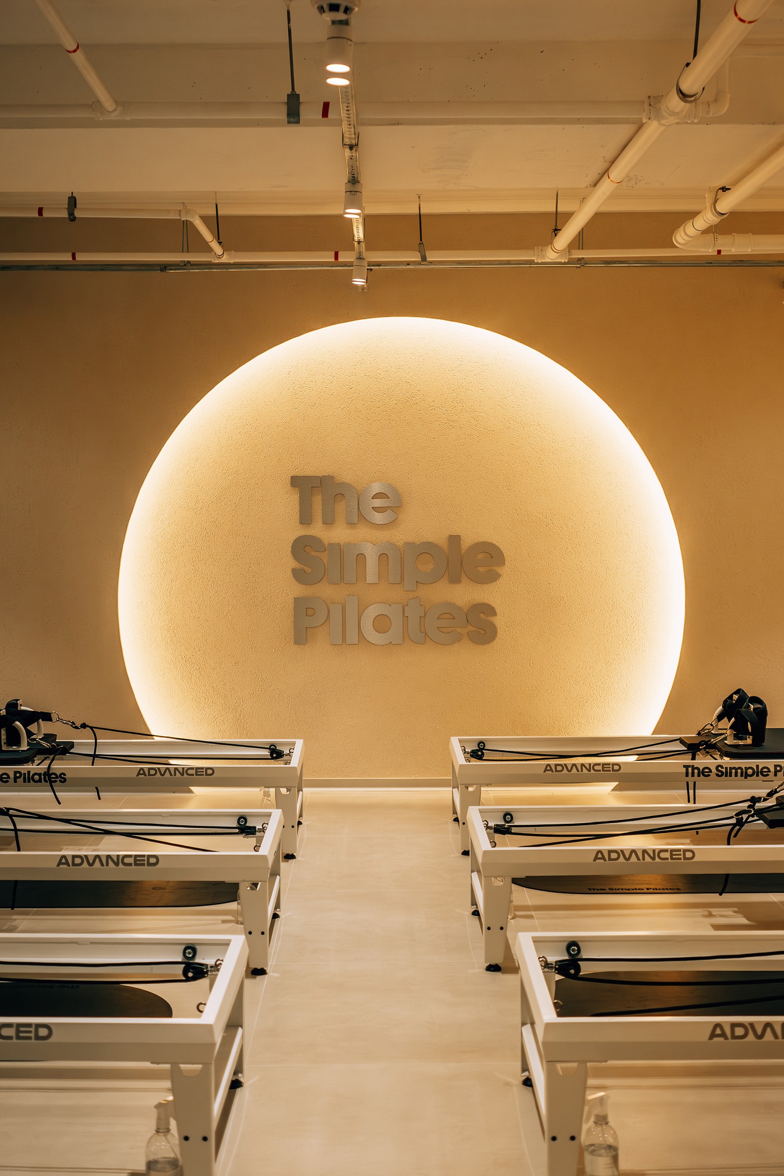Estúdio de Pilates moderno com paredes em tom bege claro e teto branco. No centro, uma parede com um círculo iluminado por trás, onde se lê The Simple Pilates em letras metálicas. Seis máquinas de Pilates brancas com detalhes pretos e a palavra ADVANCED em destaque estão dispostas em duas fileiras, três de cada lado, no chão de azulejos claros. Garrafas de água e borrifadores de limpeza são visíveis ao lado das máquinas.