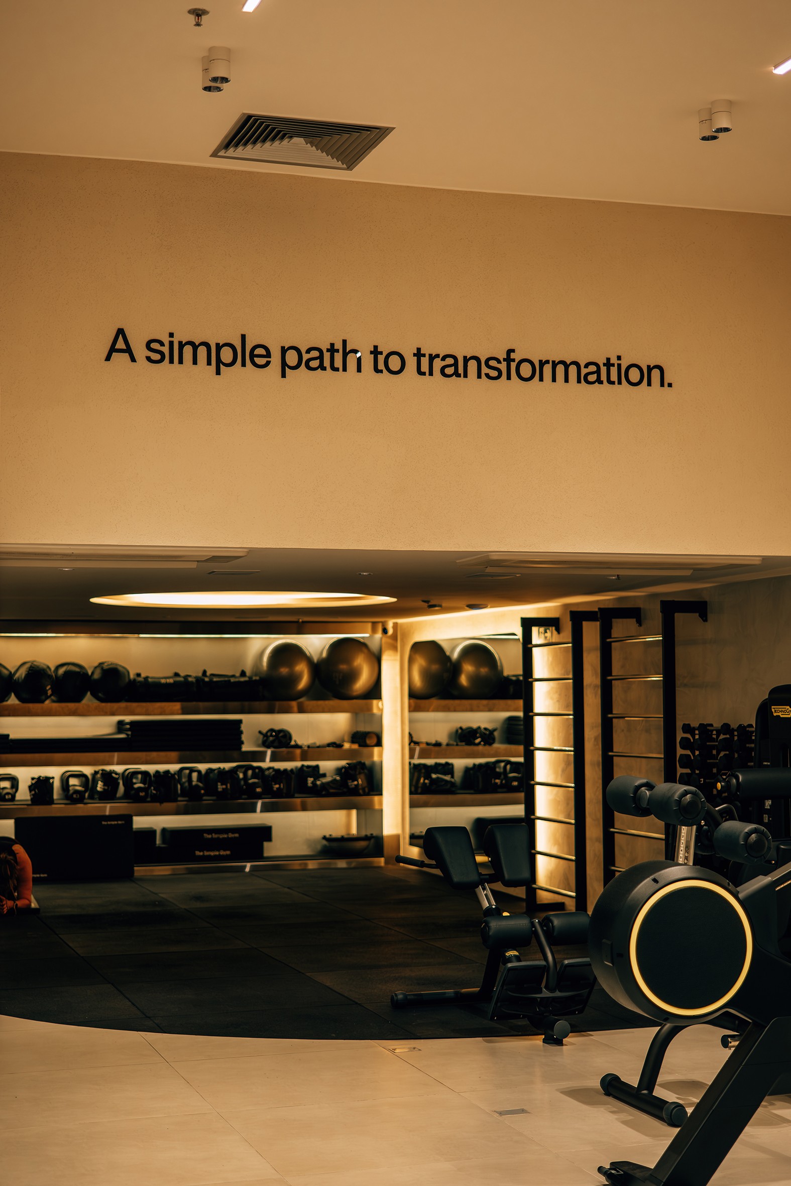 Academia moderna com piso de borracha e azulejos claros, prateleiras com halteres, kettlebells e bolas de ginástica. Na parede bege, a frase A simple path to transformation em preto. Equipamentos de musculação escuros com detalhes dourados estão à direita
