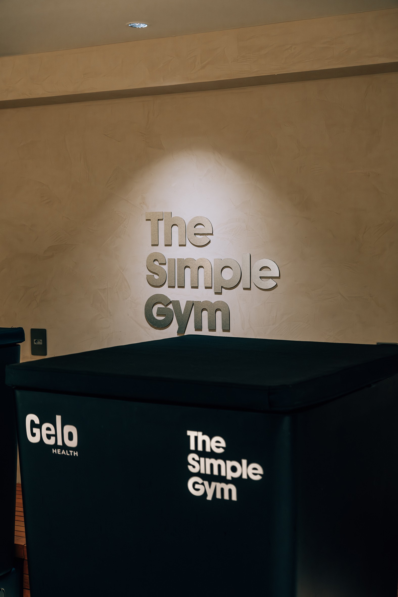 O letreiro The Simple Gym em relevo e iluminado na parede bege texturizada, acima de um balcão preto com os logotipos Gelo HEALTH e The Simple Gym em branco
