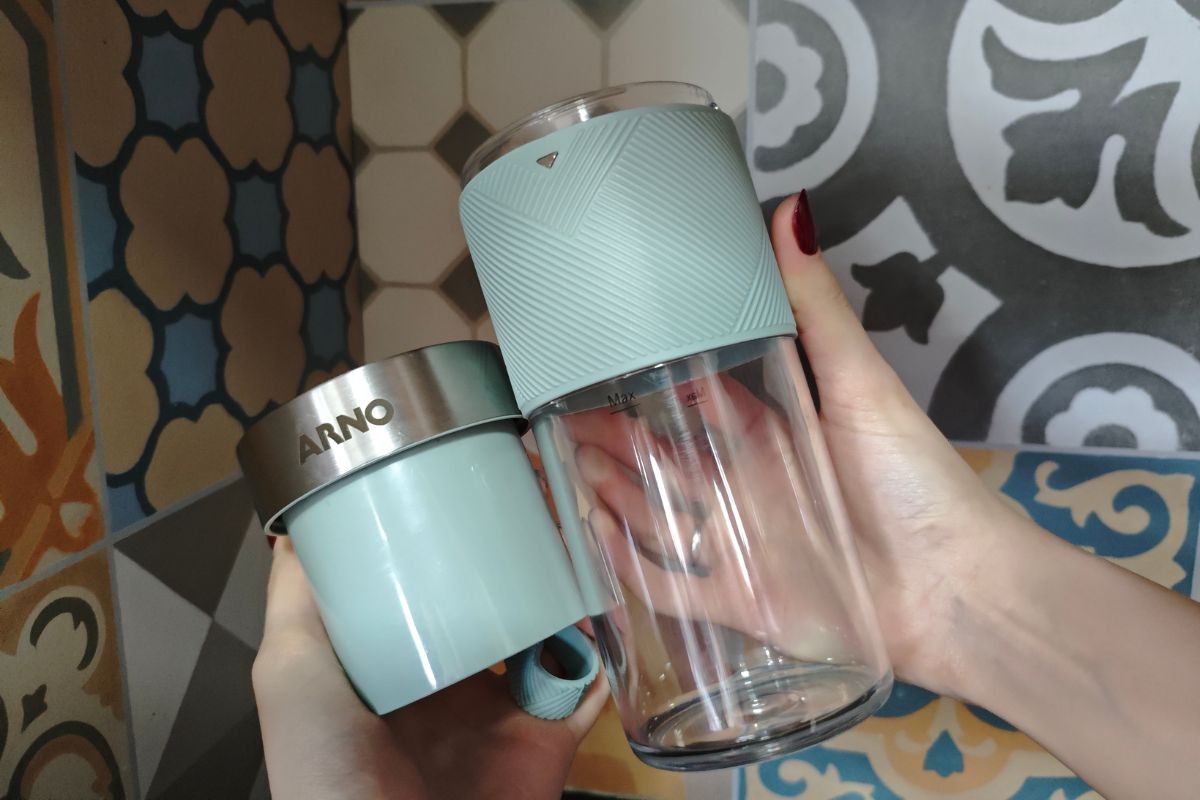 Duas m&atilde;os seguram um copo de liquidificador port&aacute;til verde-&aacute;gua e transparente com a marca Arno, sobre um fundo de azulejos coloridos