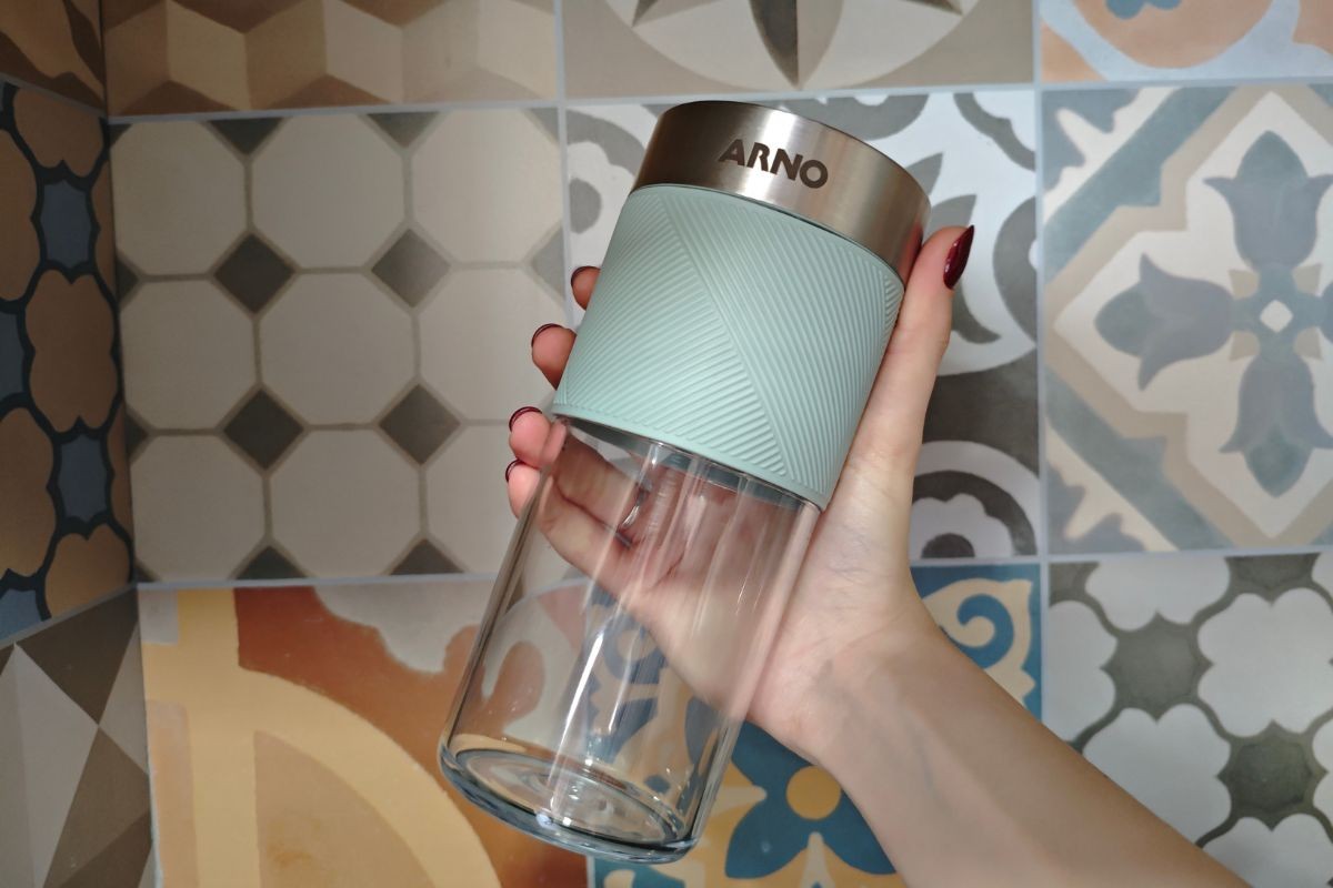 M&atilde;o feminina com unhas vermelhas segurando um copo transparente com faixa verde-&aacute;gua texturizada e tampa met&aacute;lica com o logo ARNO, sobre um fundo de azulejos coloridos com padr&otilde;es geom&eacute;tricos e florais