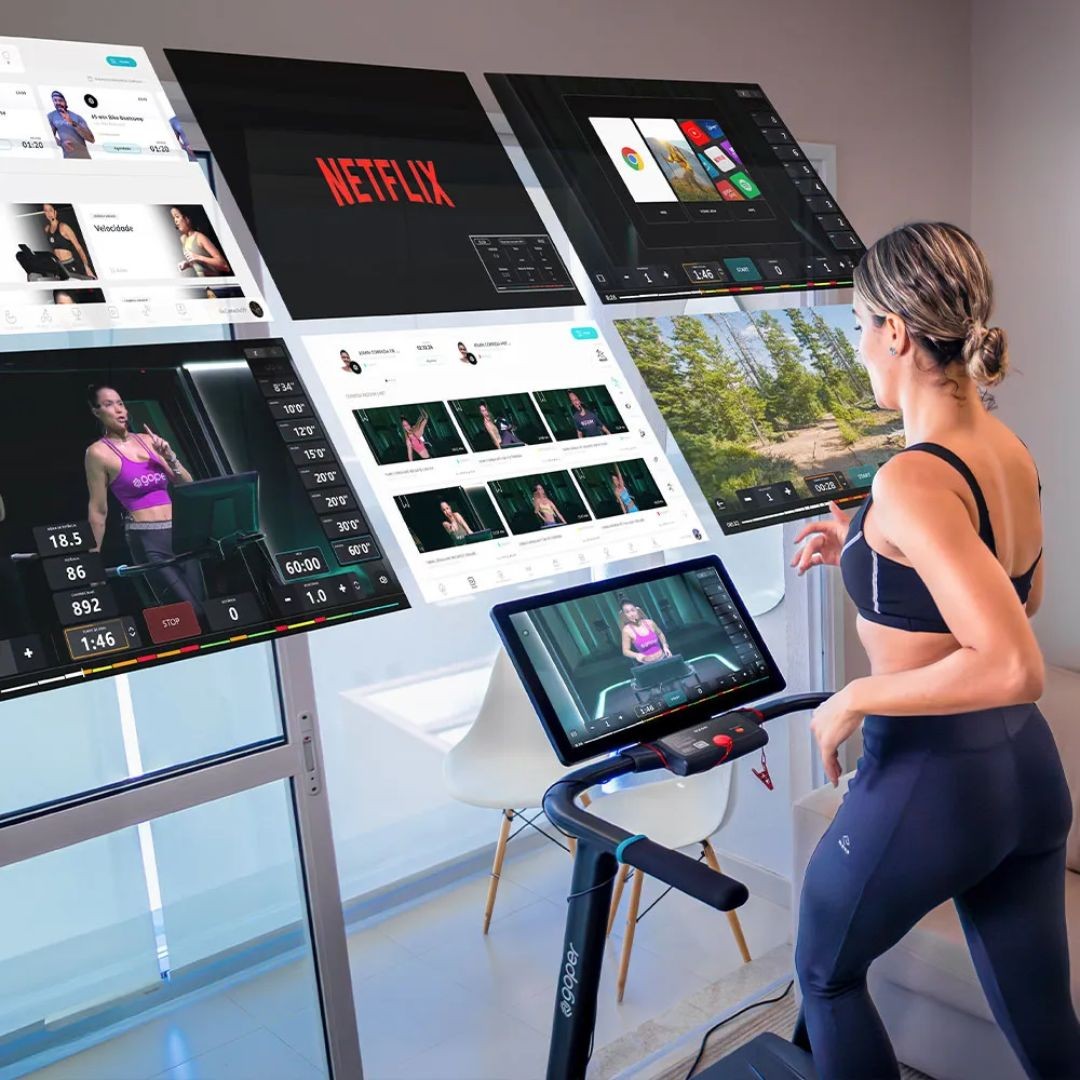 Mulher de costas, com cabelo preso e roupa de ginástica preta, correndo em esteira moderna com tela que exibe uma aula. Ao redor, várias telas flutuantes mostram aplicativos de treino, Netflix e paisagens, sugerindo opções de entretenimento e exercícios