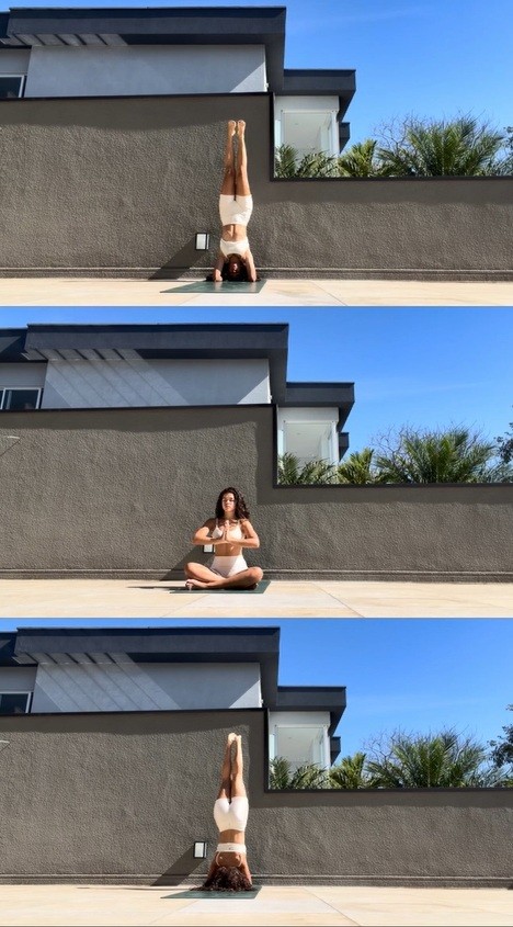 Mulher de cabelo cacheado escuro, top e shorts brancos, praticando yoga em três poses: invertida, sentada com mãos em prece, e invertida novamente, sobre um tapete verde em um terraço com parede cinza e céu azul
