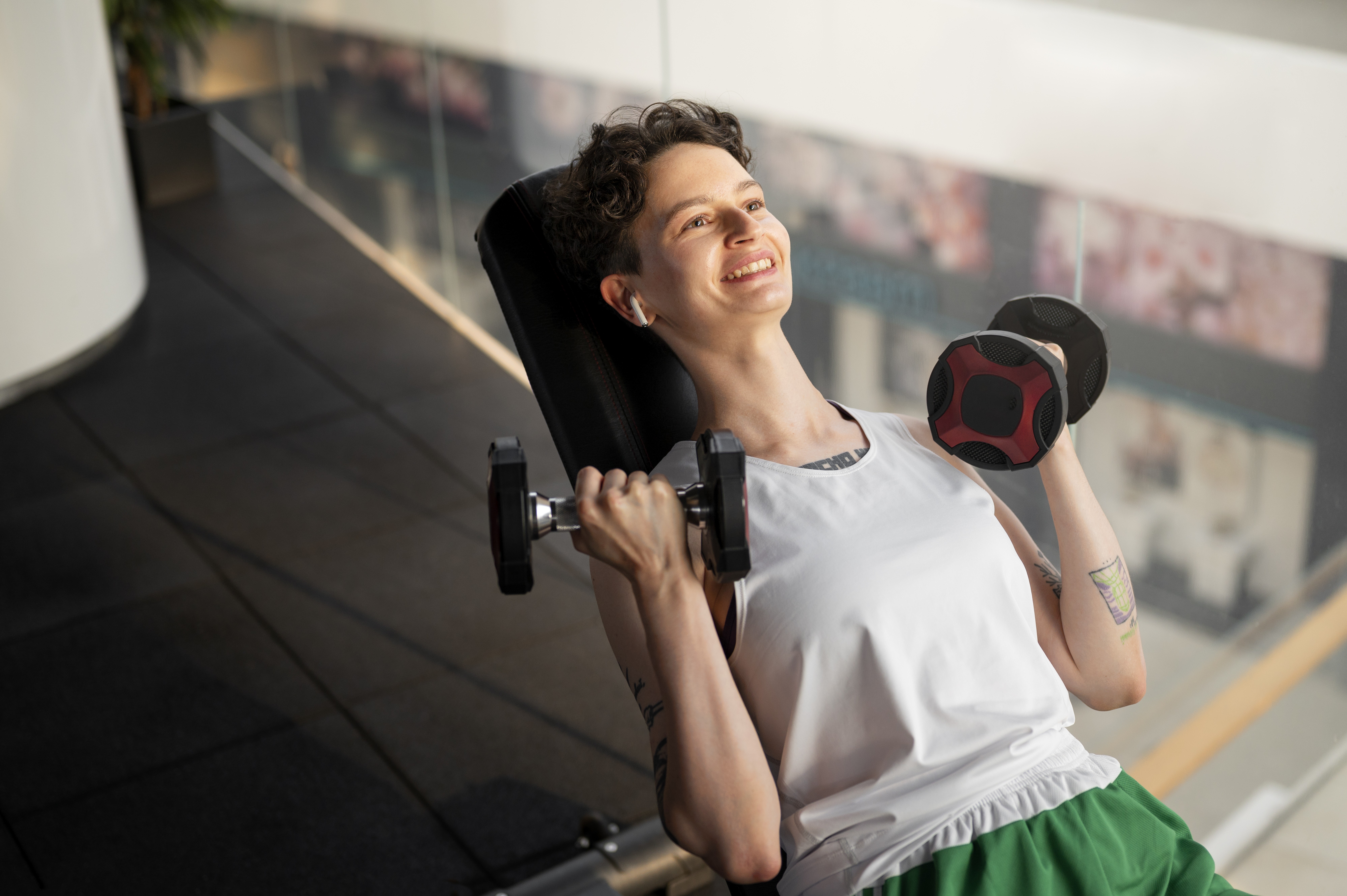 Mulher de cabelo curto e encaracolado, sorrindo e olhando para cima, fazendo exercícios com halteres em um banco de academia. Ela usa fones de ouvido brancos, regata branca e shorts verdes, exibindo tatuagens nos braços. O fundo é um espaço amplo e moderno com piso escuro e reflexos