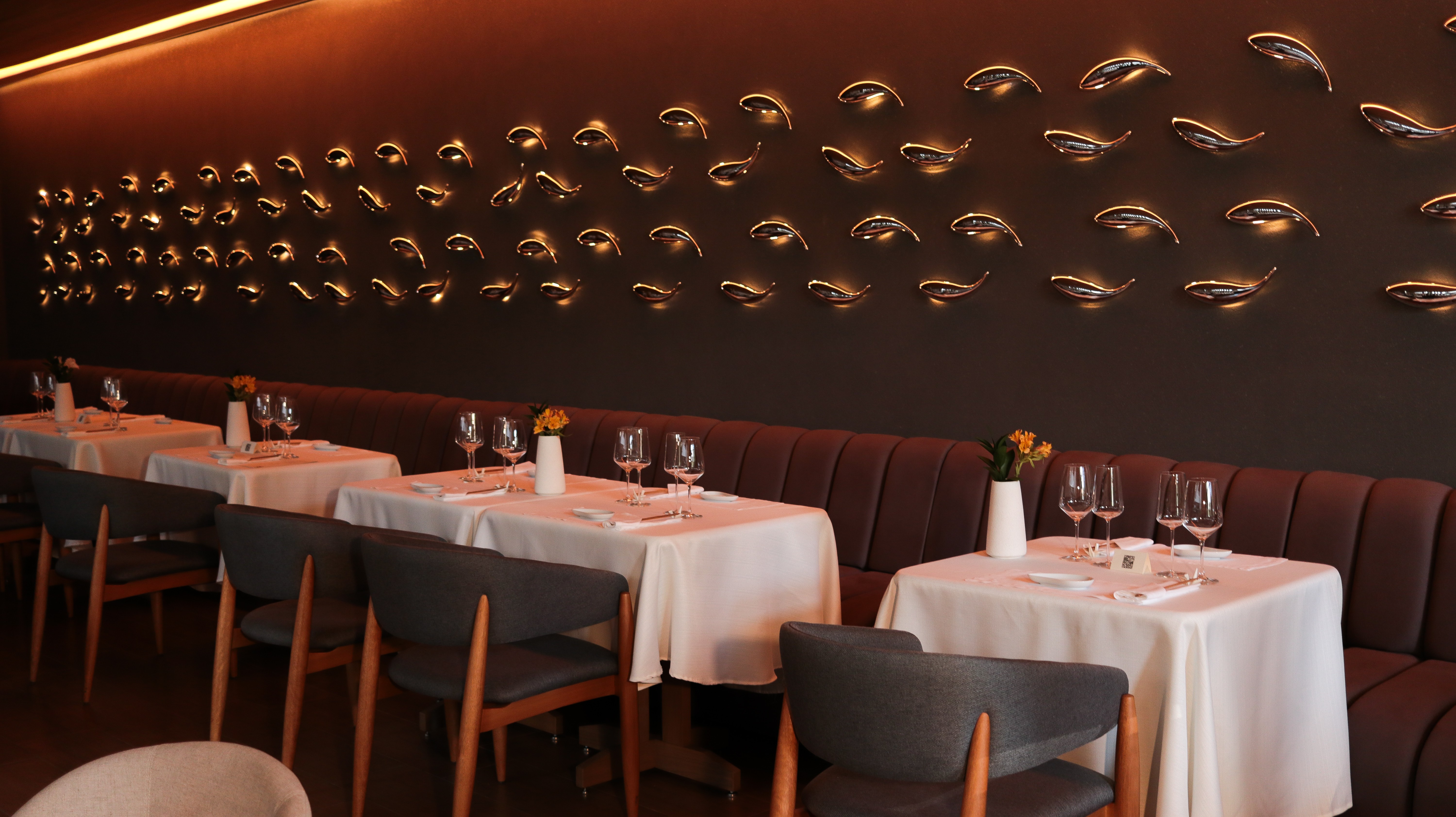 Salão de restaurante elegante com mesas postas, cadeiras de madeira e estofamento cinza, e um longo sofá marrom. A parede escura é decorada com dezenas de luminárias em formato de peixe, emitindo luz dourada