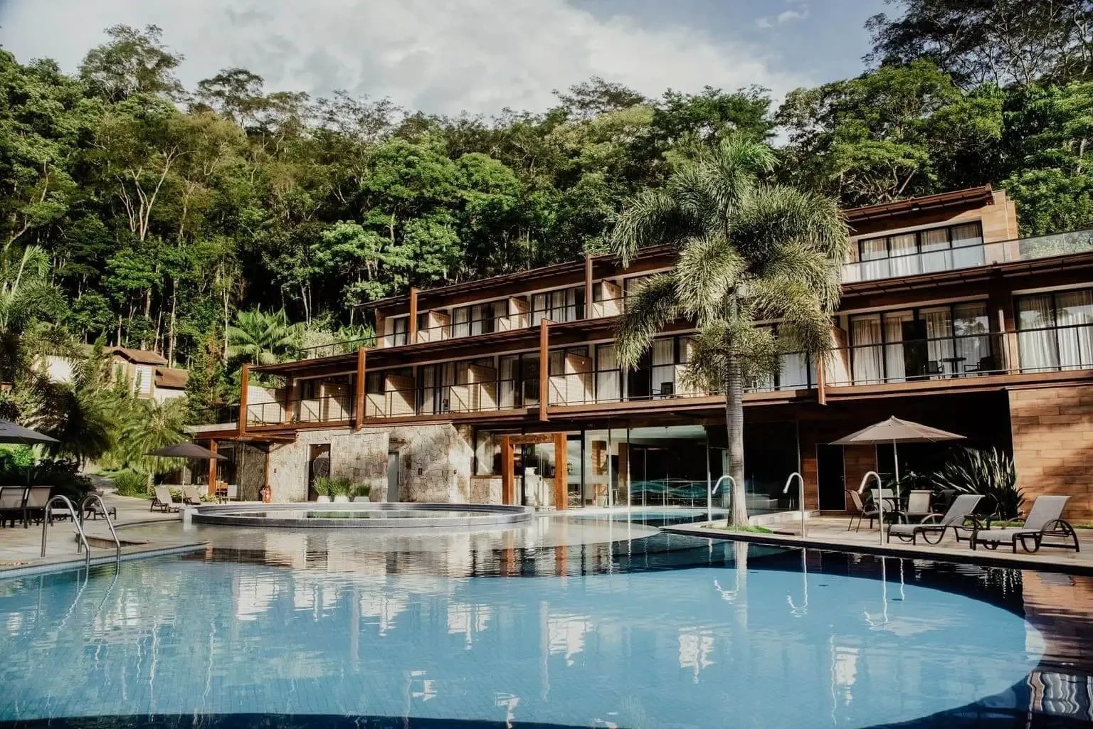 Hotel moderno com fachada de madeira e pedra, varandas com grades pretas, piscina azul com borda infinita e espreguiçadeiras, palmeiras e vegetação exuberante ao fundo sob céu nublado