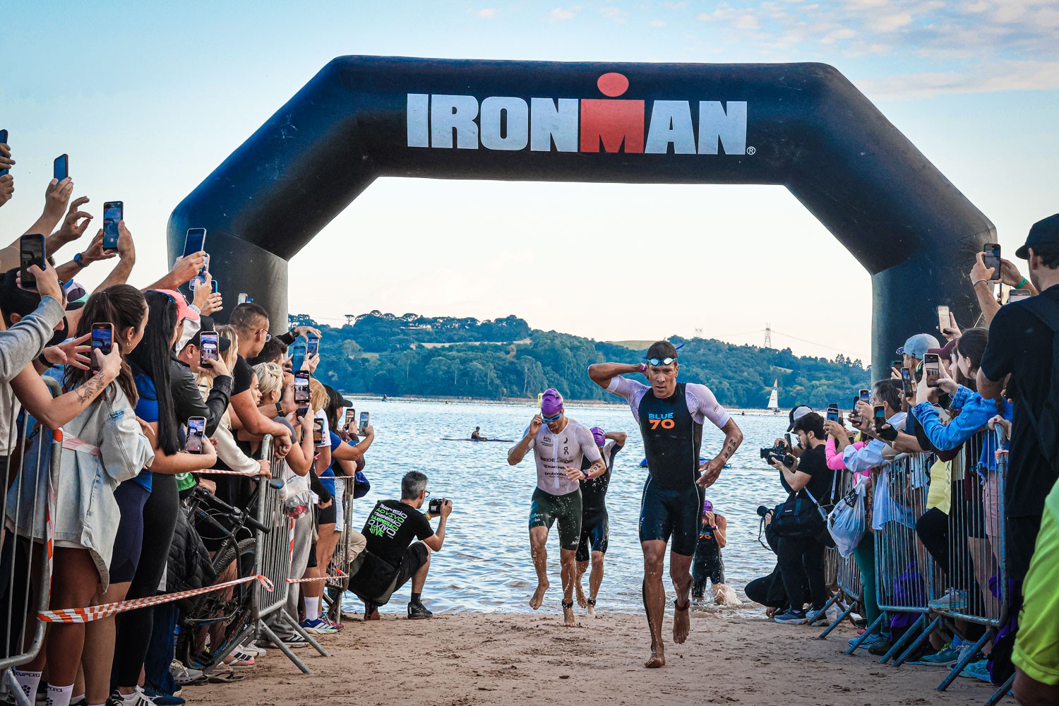 Atletas de triathlon saem da água e correm na areia sob um arco inflável preto com o logo IRONMAN. Uma multidão de espectadores filma com celulares, enquanto um fotógrafo agachado registra a cena à beira do lago.