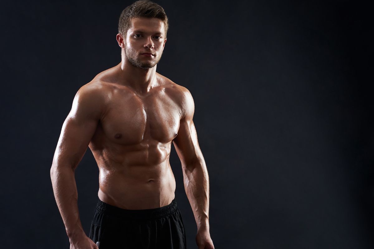 Homem musculoso, sem camisa, com suor brilhando na pele, olhando diretamente para frente, em um fundo escuro