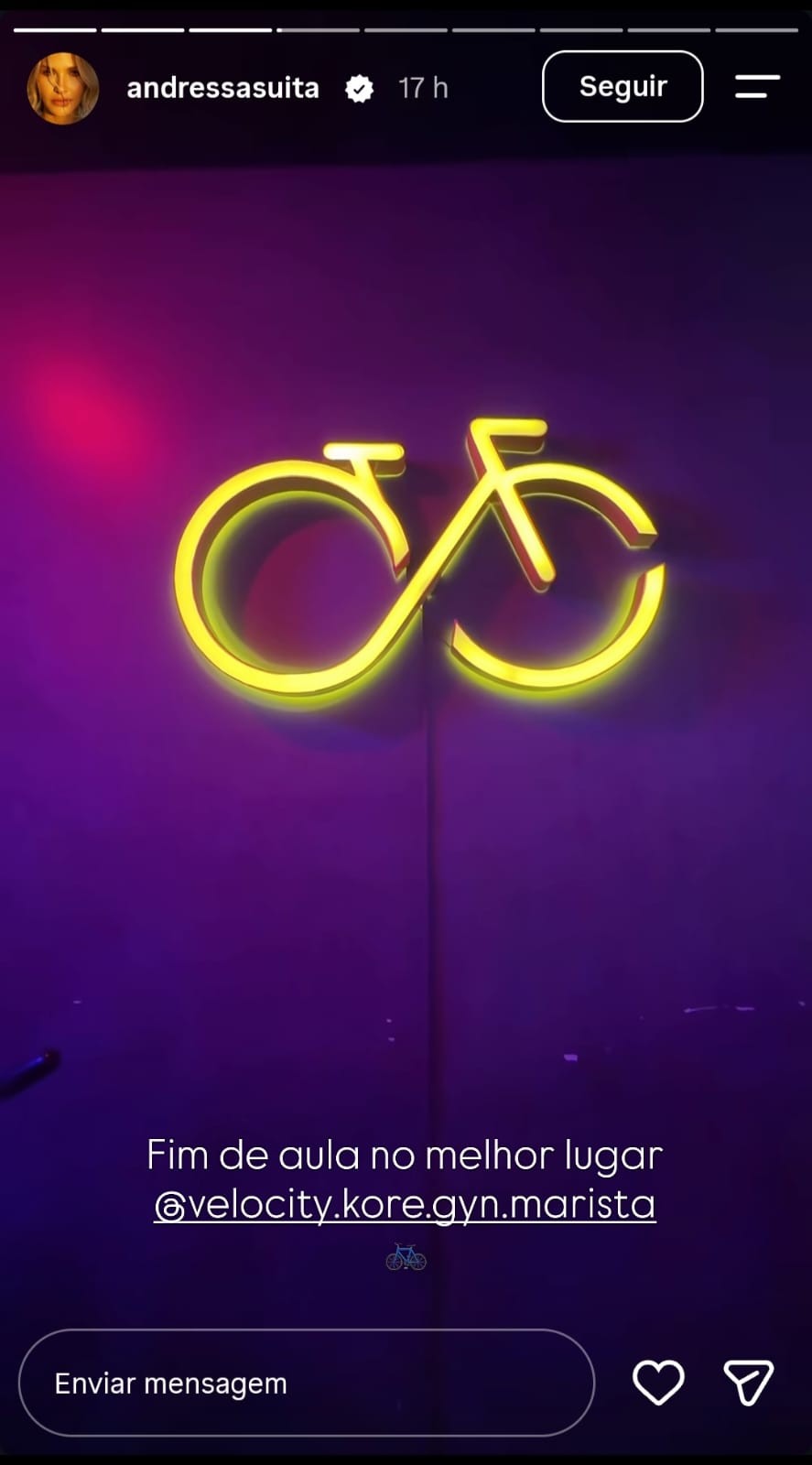 Logotipo de bicicleta em neon amarelo brilhante sobre uma parede roxa escura, com reflexos de luz rosa e azul. Abaixo, texto branco Fim de aula no melhor lugar @velocity.kore.gyn.marista
